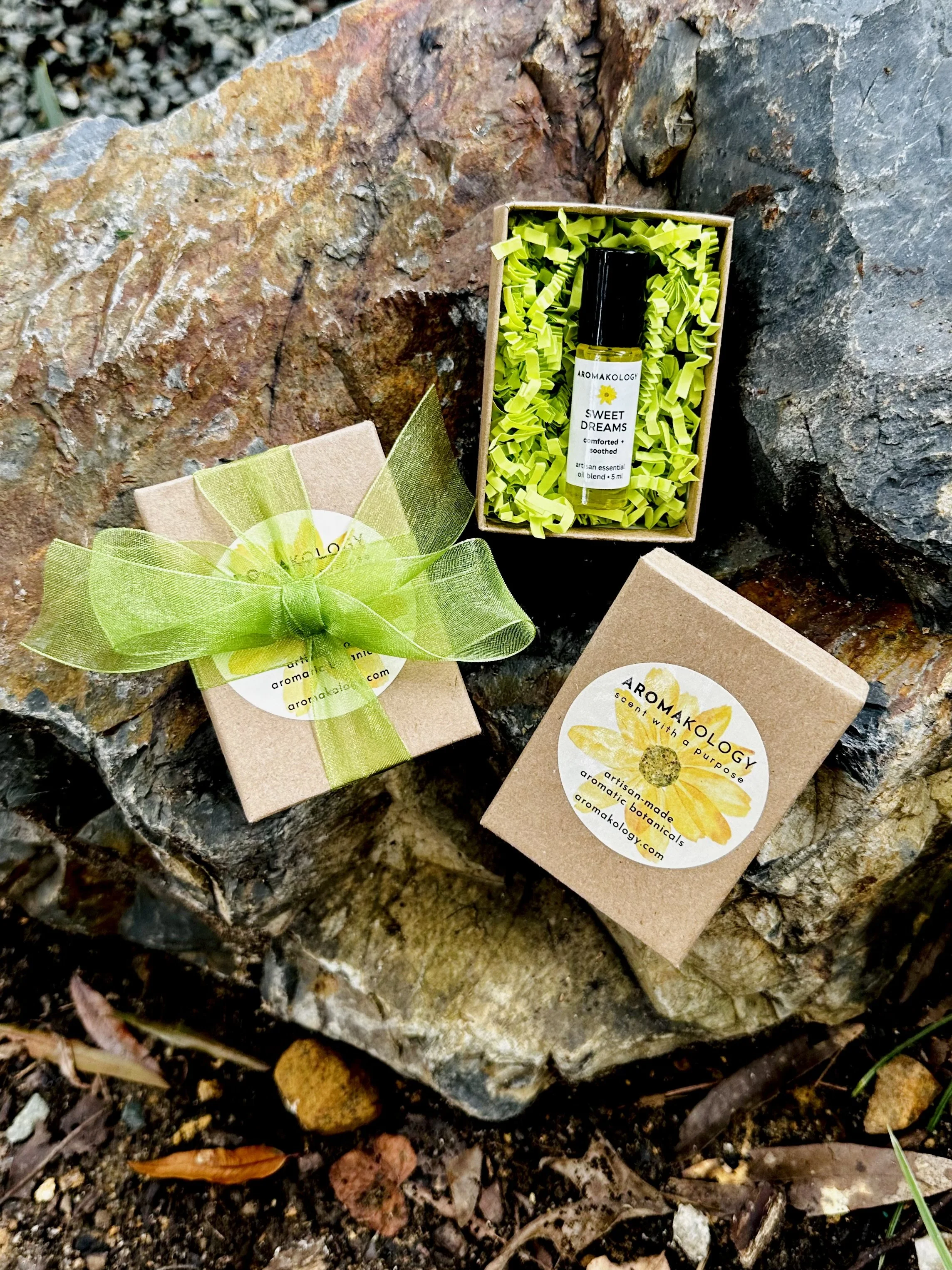 A small gift box with a green ribbon, a set of aromatic essential oils, and a cardboard box with flower branding, all placed on a large rock outdoors.