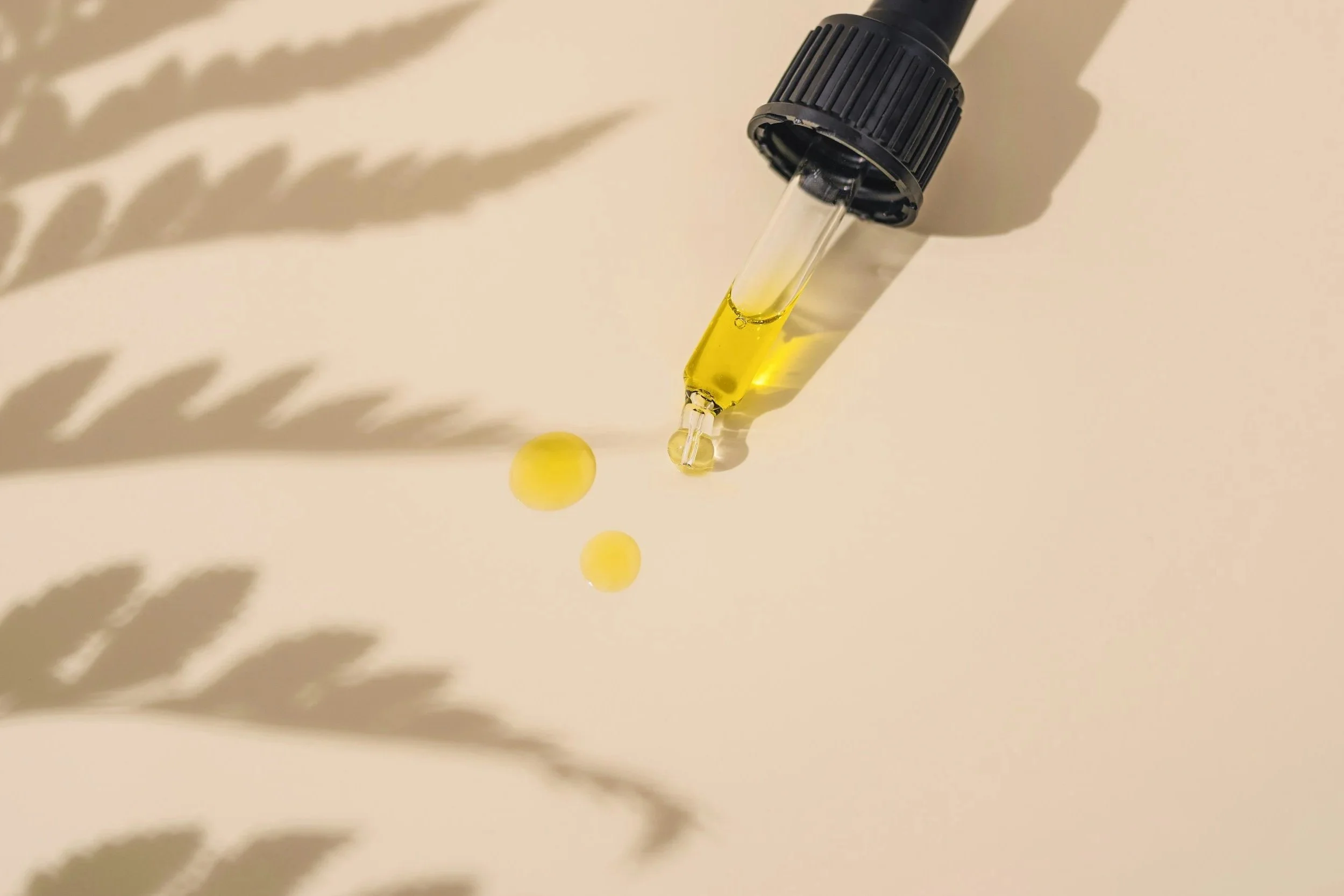 A glass dropper bottle with yellow oil and two yellow capsules on a beige surface, casting shadows.