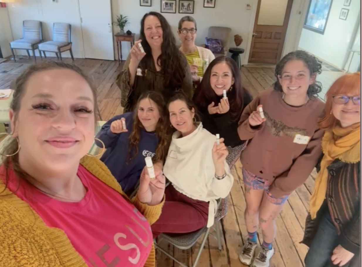 A group of eight women smiling and holding small bottles, gathered in a cozy indoor space with wooden floors and art on the walls.