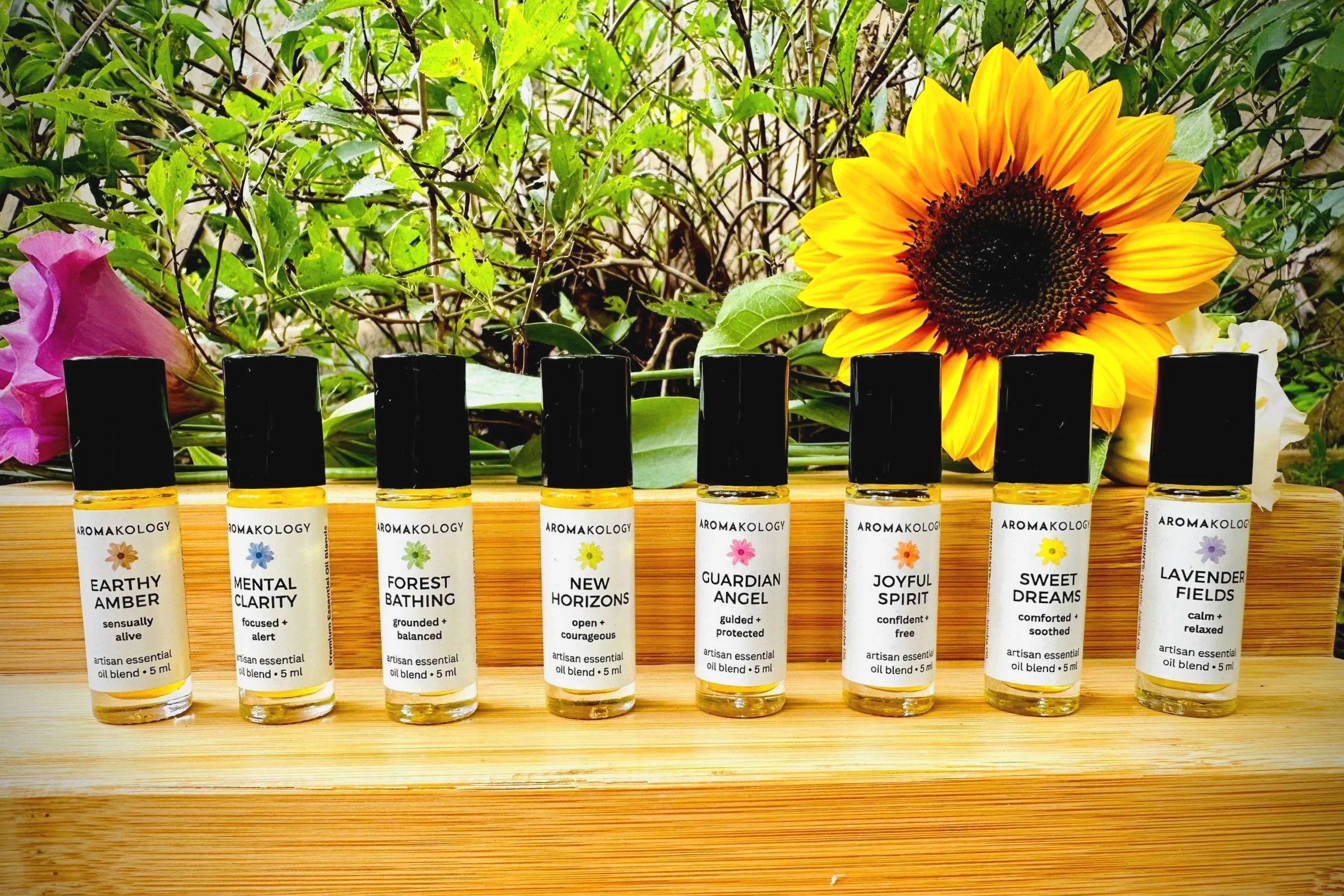 A row of nine essential oil bottles labeled with different names and descriptions, placed on a wooden surface with a garden and sunflowers in the background.