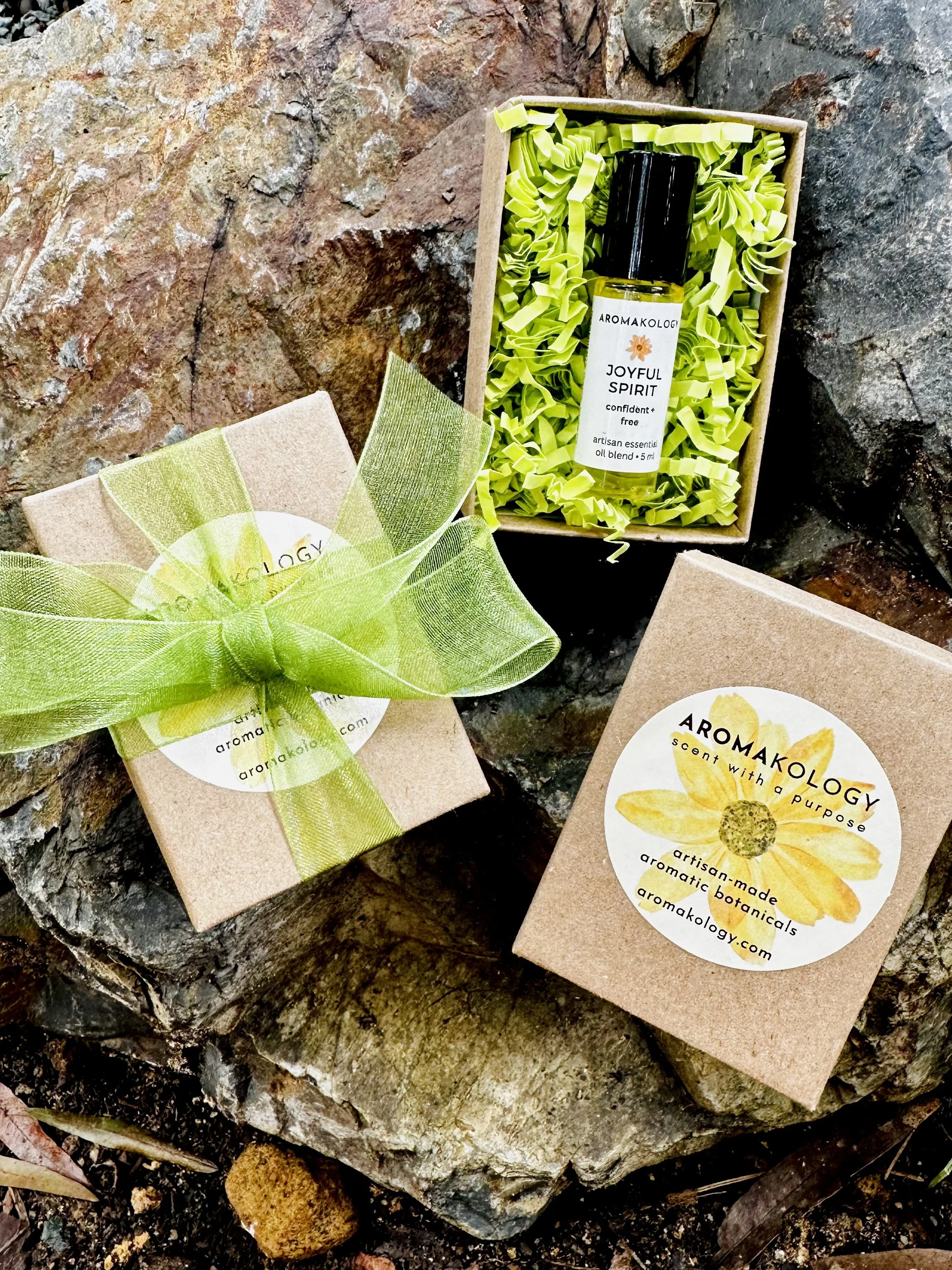 A small gift box wrapped with a light green ribbon, a cardboard box with airflow paper and a small essential oil bottle inside, and a cardboard box with a yellow flower label, all placed on rocks.