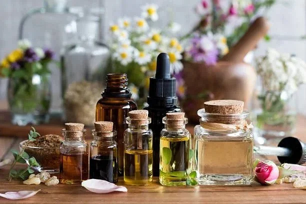 Various bottles of essential oils, a dropper, flowers, and herbs on a wooden surface with a blurred background of more flowers and plants.