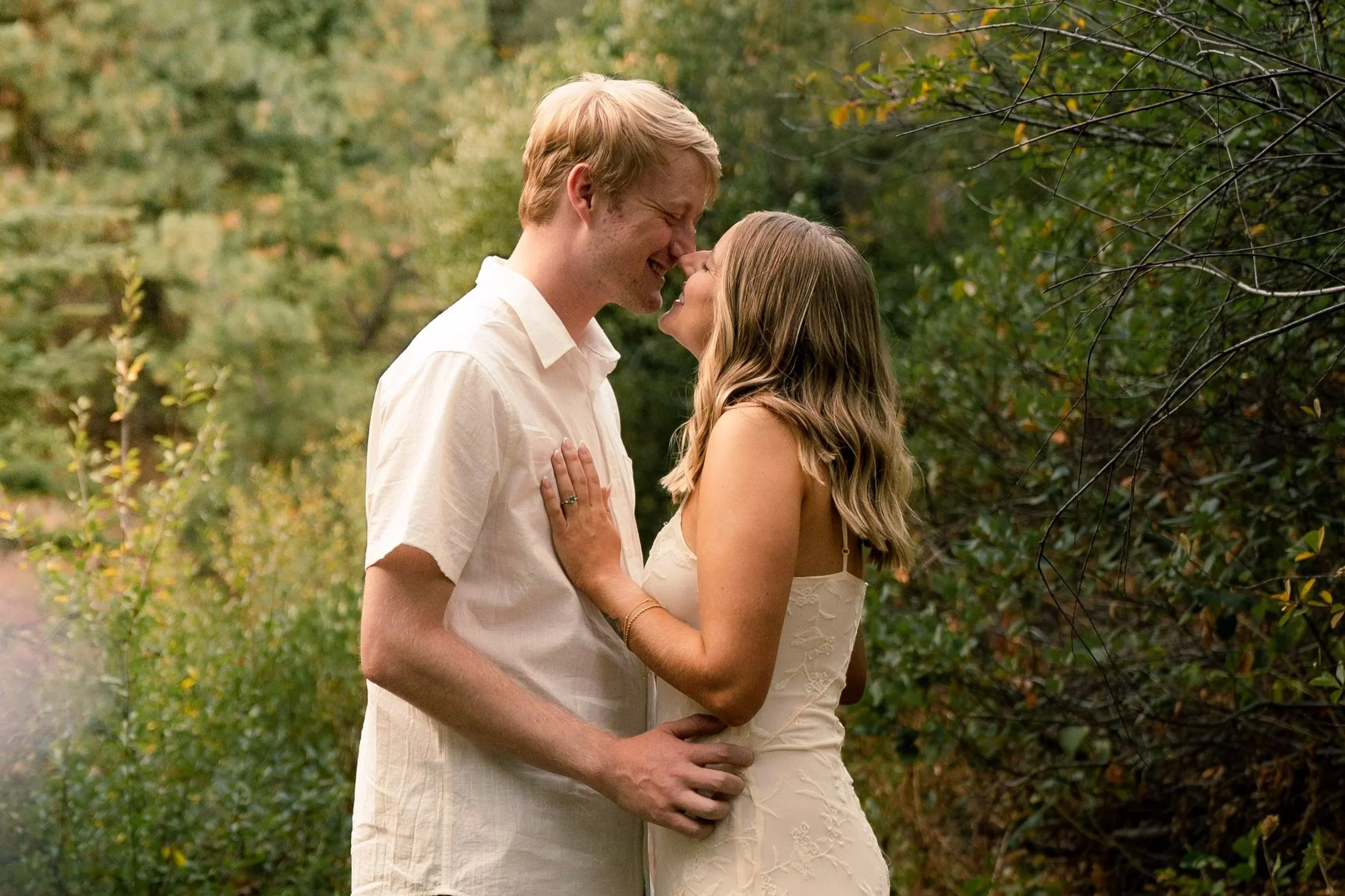 A young couple stands close together outdoors in a lush green forest, smiling and touching noses in an intimate moment.