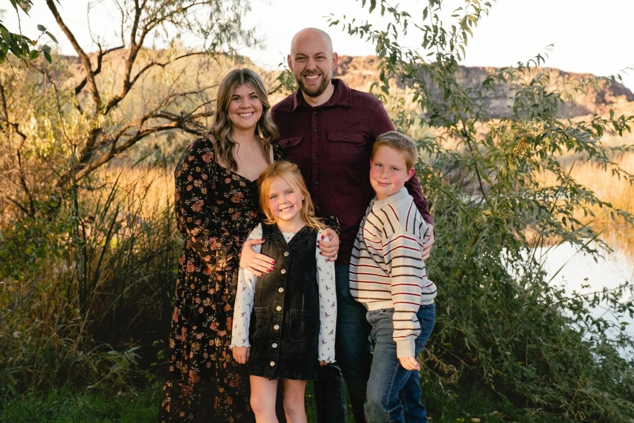 A family of four smiling outdoors near a body of water with hills in the background, surrounded by trees and foliage, during sunset.