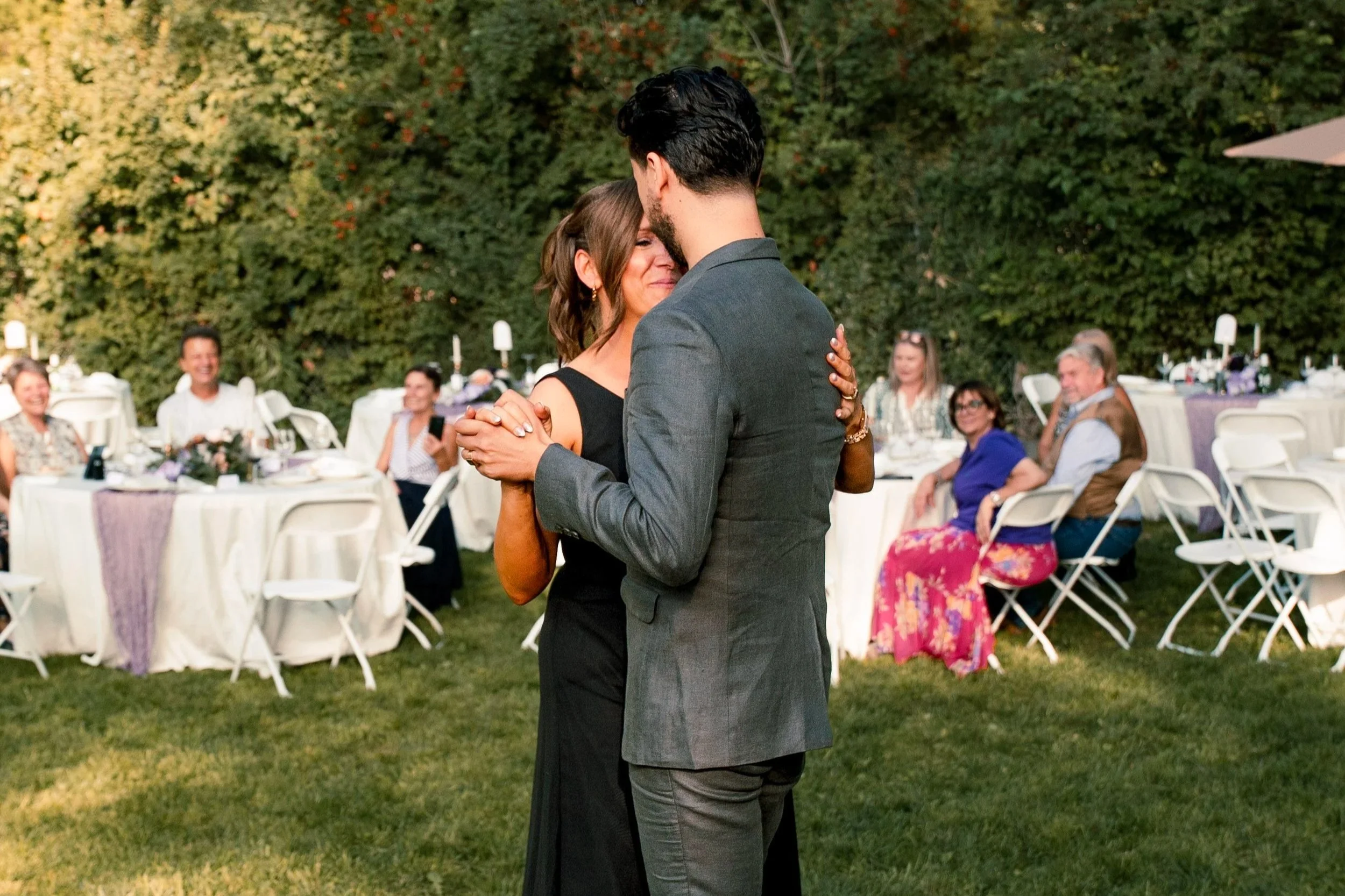 A couple dancing closely at an outdoor wedding reception, with guests seated at round tables decorated with flowers and candles, surrounded by greenery.