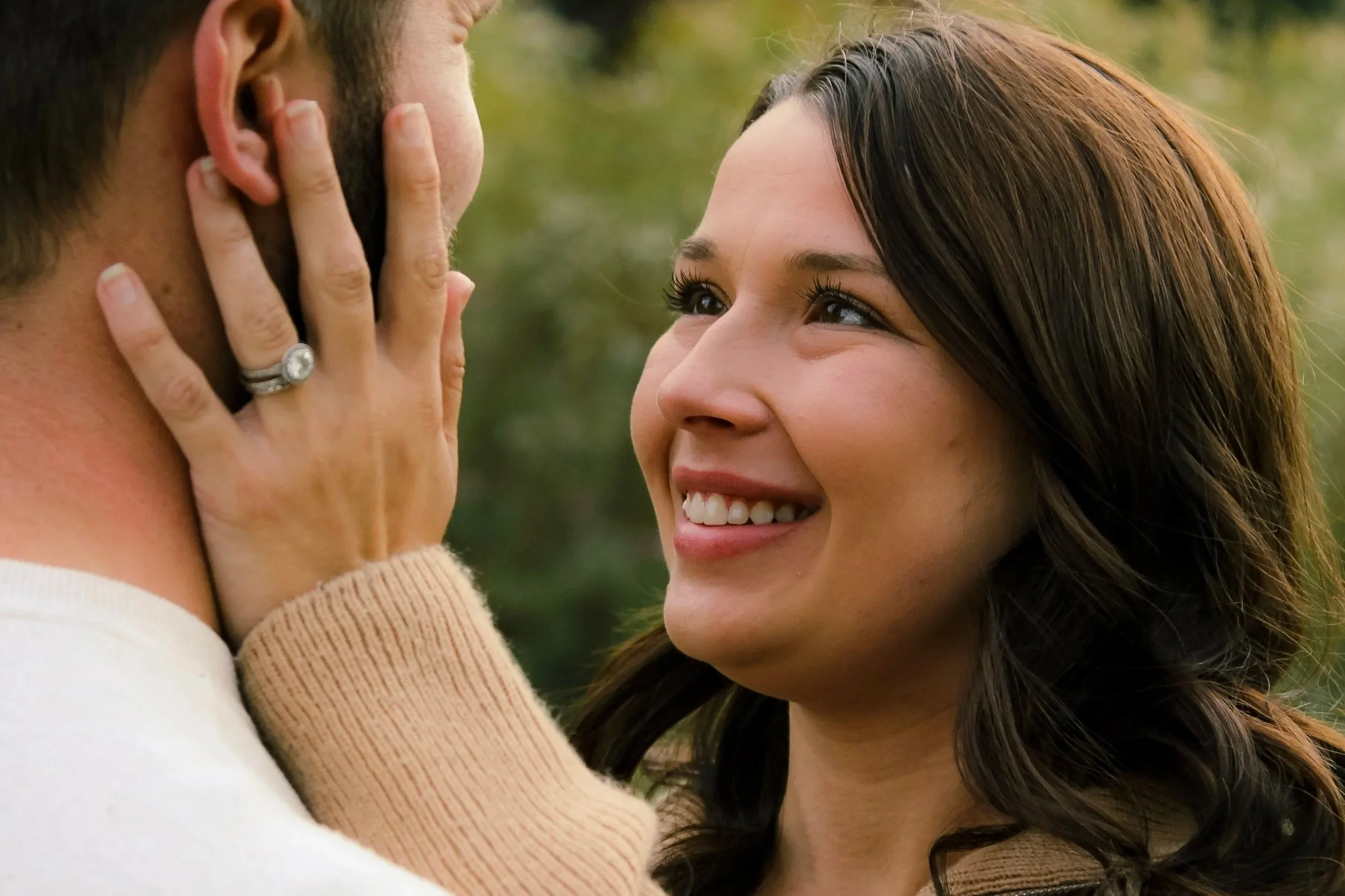 A woman with brown hair and a bright smile looks lovingly at a man with short dark hair, who is close to her face. The man's face is partially visible as he gently touches her cheek, outdoors with a blurred green background.