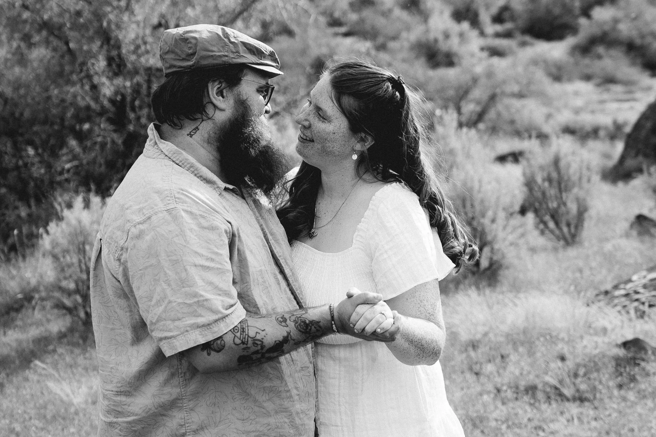 A black-and-white photo of a couple holding hands and smiling at each other outdoors, with trees and grass in the background.