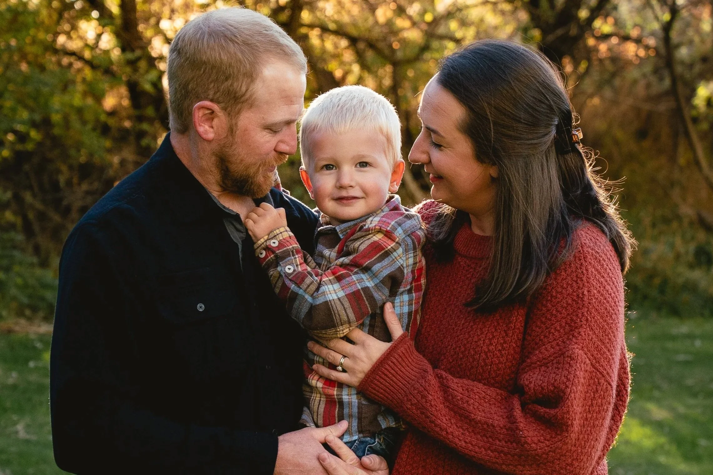 A happy family of three, a man, a woman, and a young boy, enjoying a moment outdoors during autumn with trees in the background. The man and woman are facing each other and smiling, while the boy is in the middle, looking at the camera with a slight 