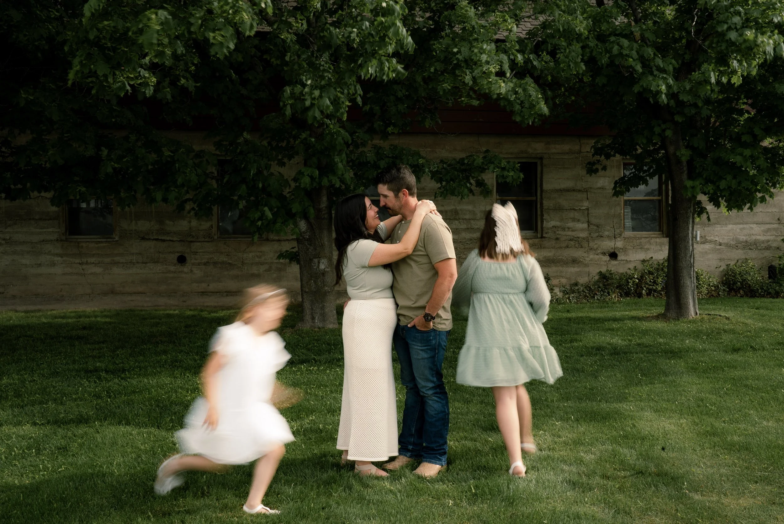 A couple hugging outdoors under green leafy trees, with two young girls in white dresses running in the foreground.