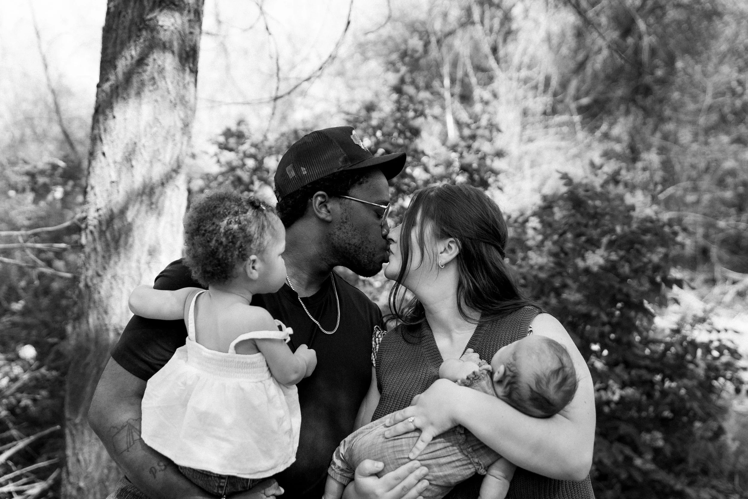 A family of five, including two children, sharing a kiss in a park with trees in the background.