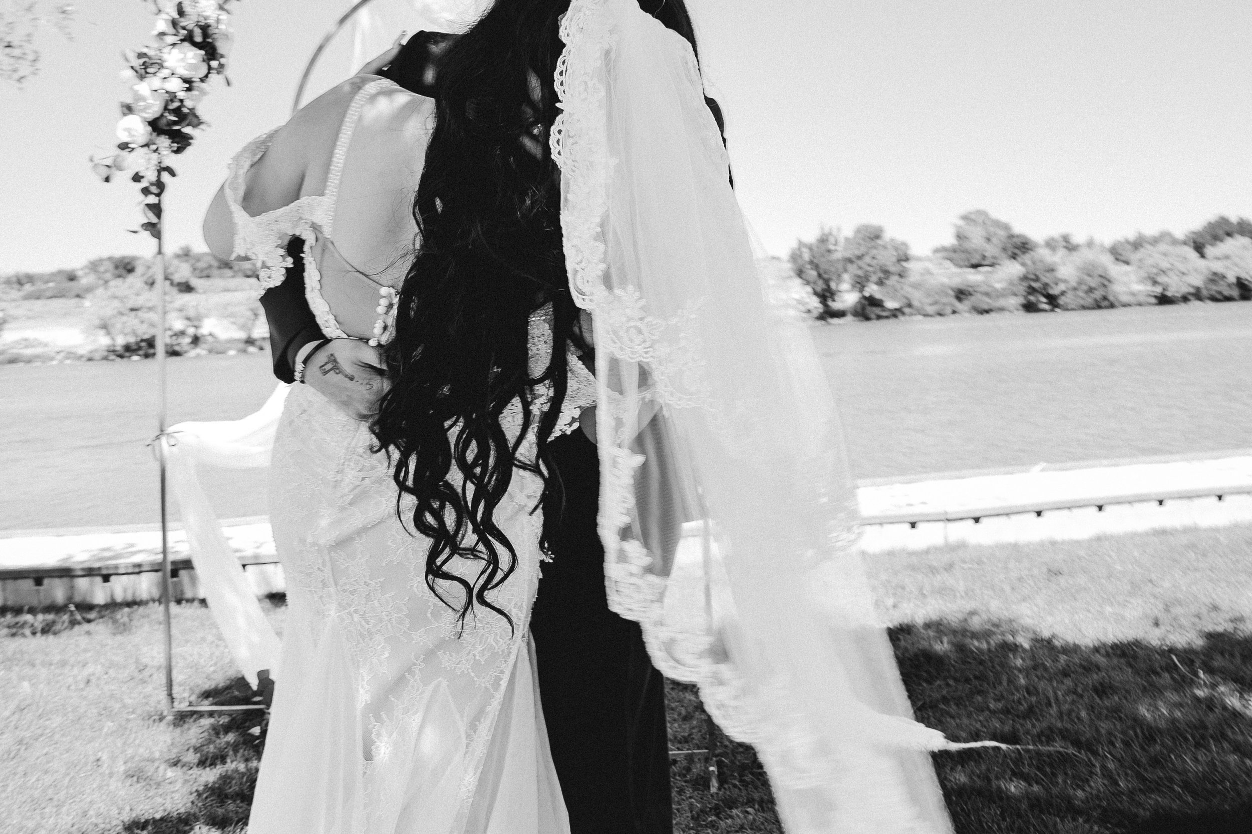 A bride with long dark wavy hair wearing a lace wedding dress and veil, standing outdoors with a groom, in a black and white photo.