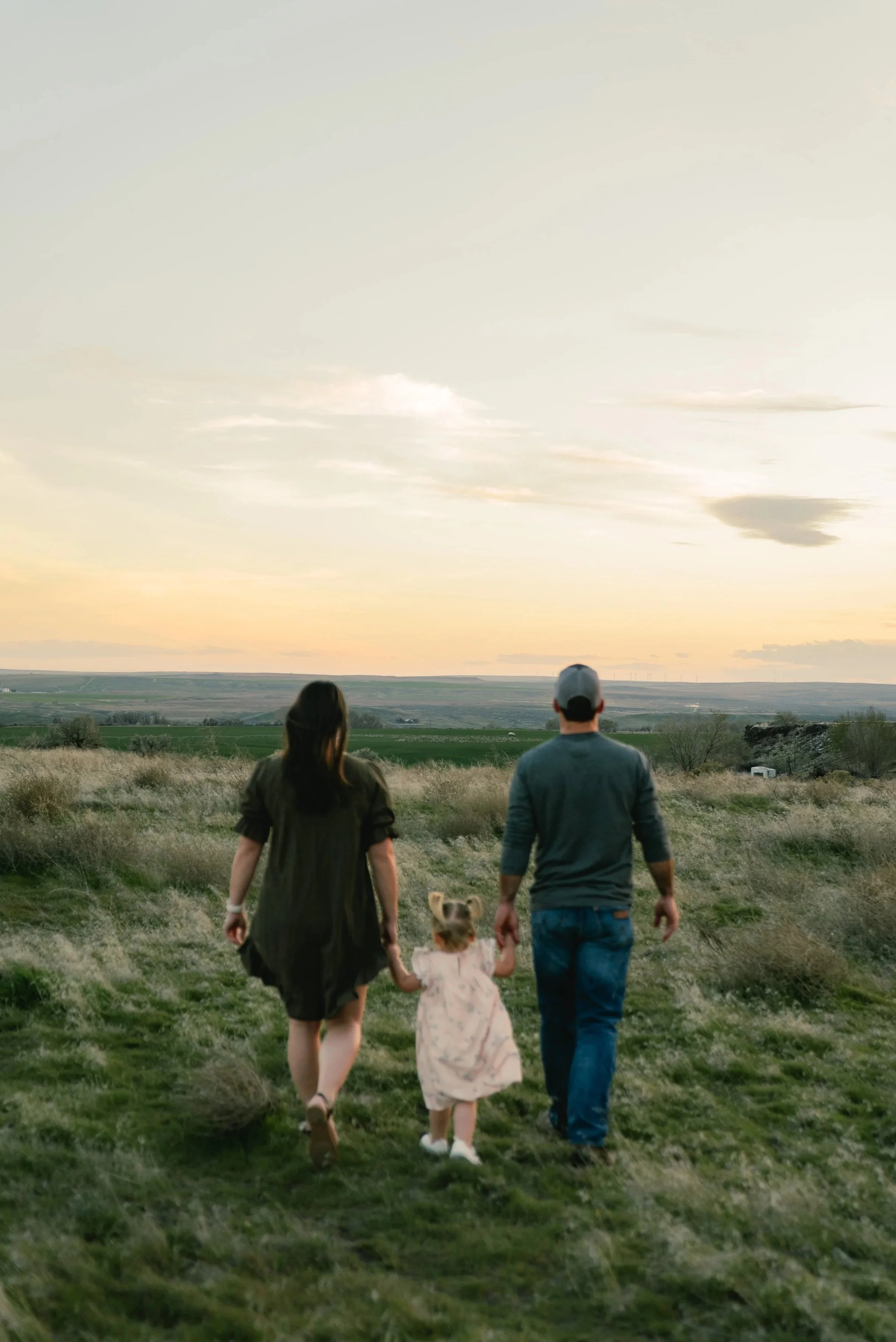 A family of three, a man, a woman, and a little girl, holding hands and walking through a grassy field toward the sunset, with open countryside in the background.