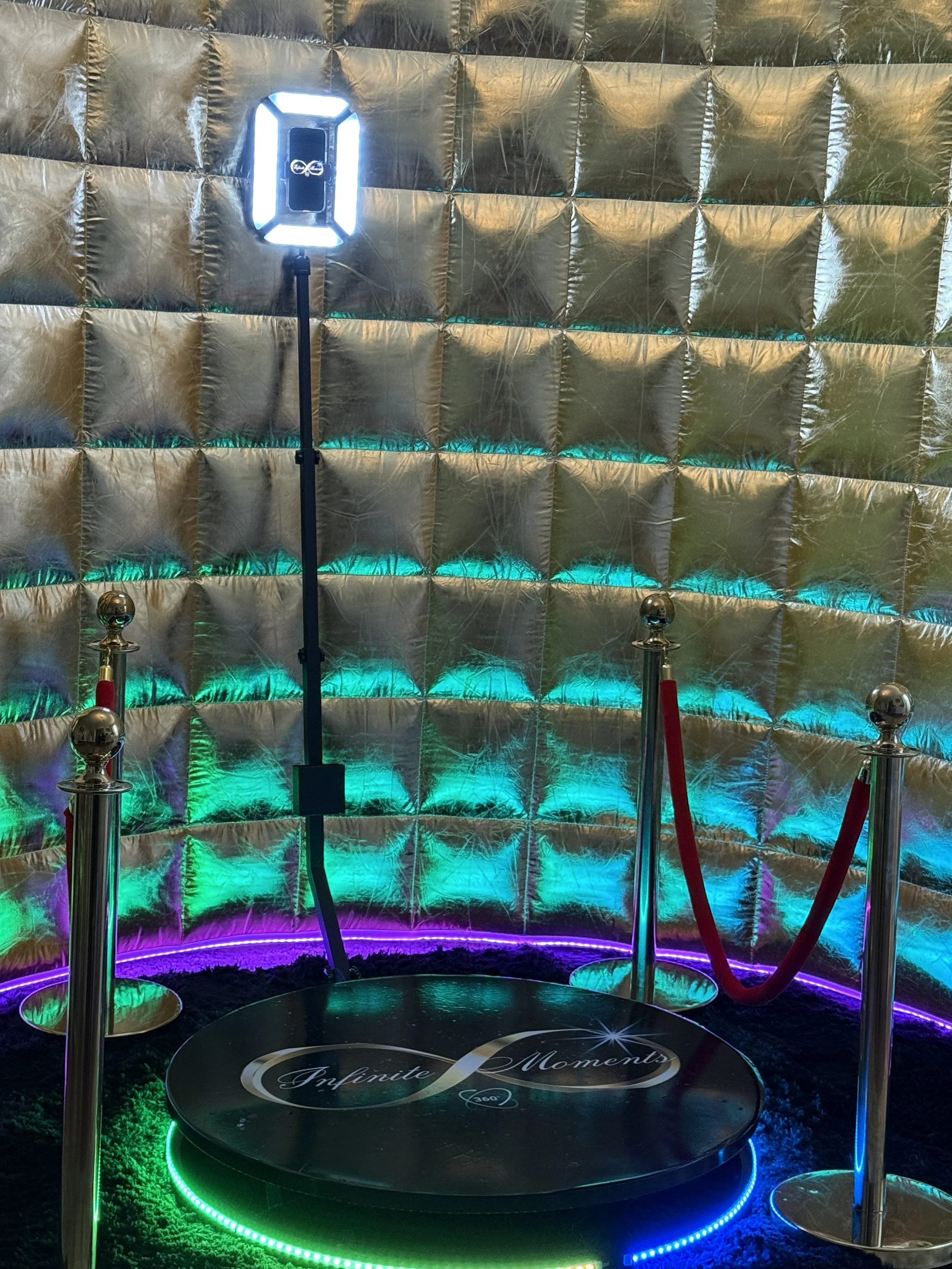 Colorful, illuminated art installation with a circular platform labeled 'Infinite Moments,' surrounded by velvet ropes, against a shiny, iridescent backdrop.