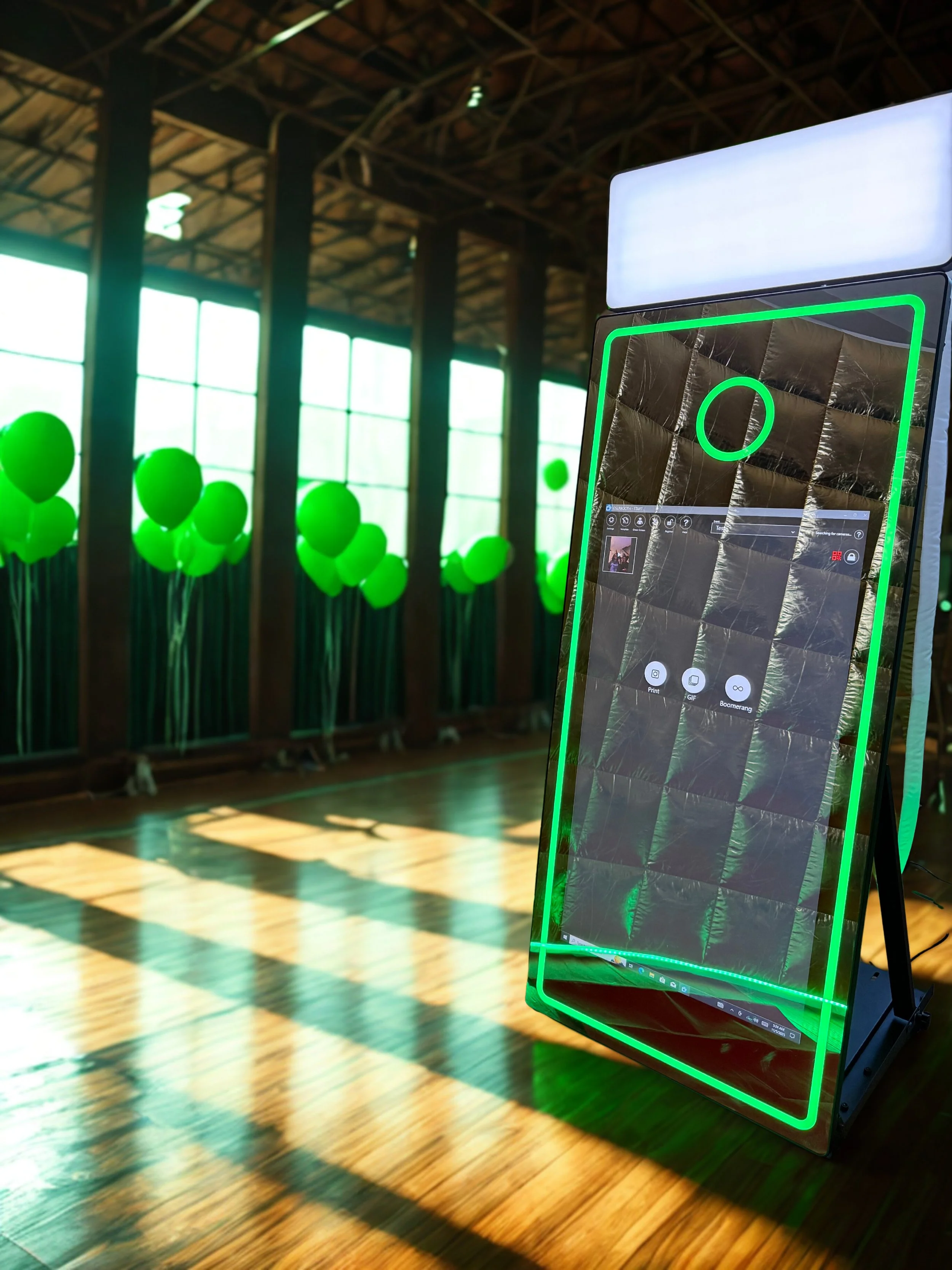 A digital photo booth with a green neon border in a room with large windows, green balloons, and wooden floors.