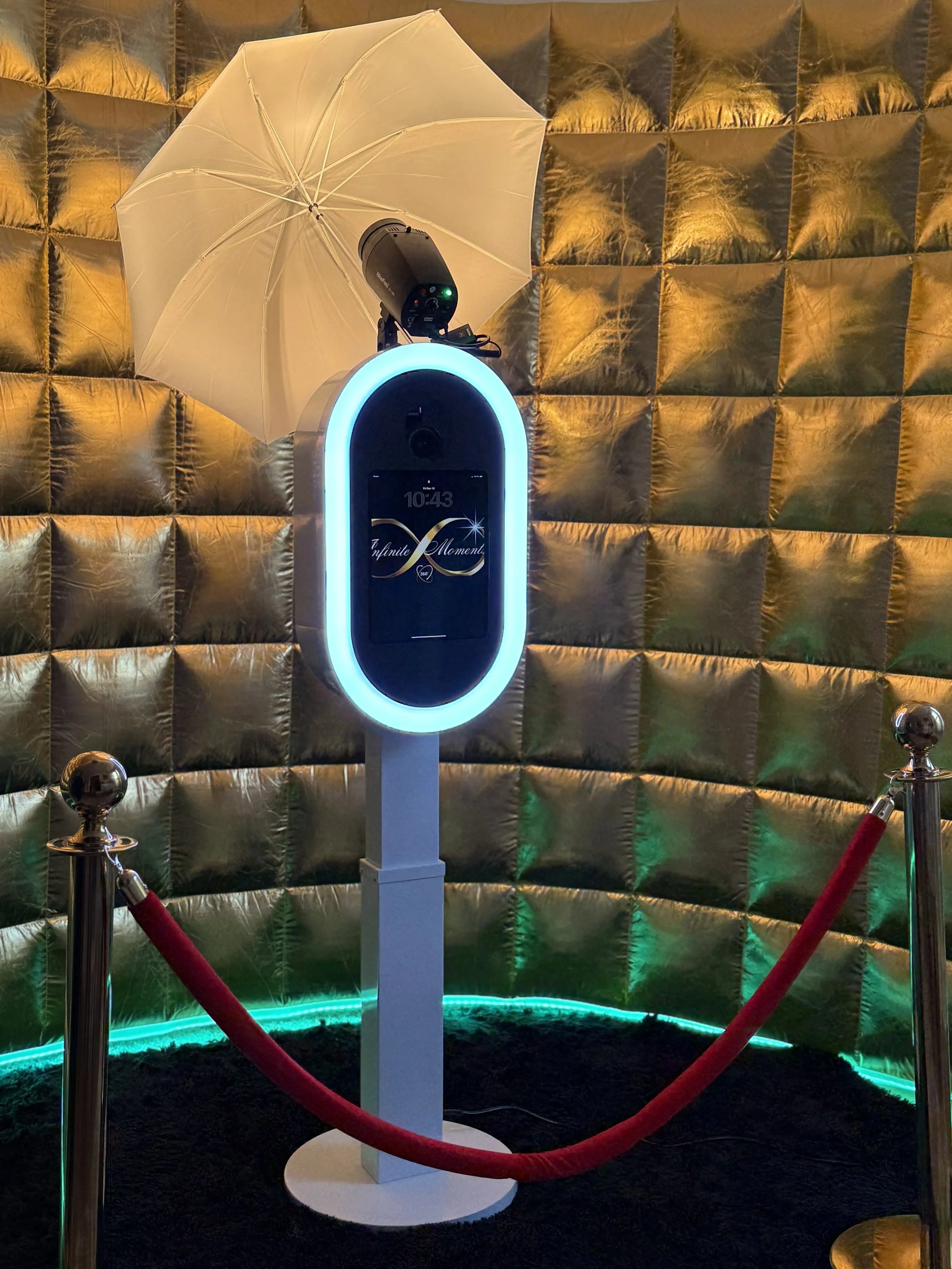 A high-tech kiosk with a touchscreen display, illuminated with blue LED lights, is surrounded by red velvet ropes. Behind it, there is a golden quilted backdrop, and an umbrella with a light-colored canopy is mounted on top of the kiosk.