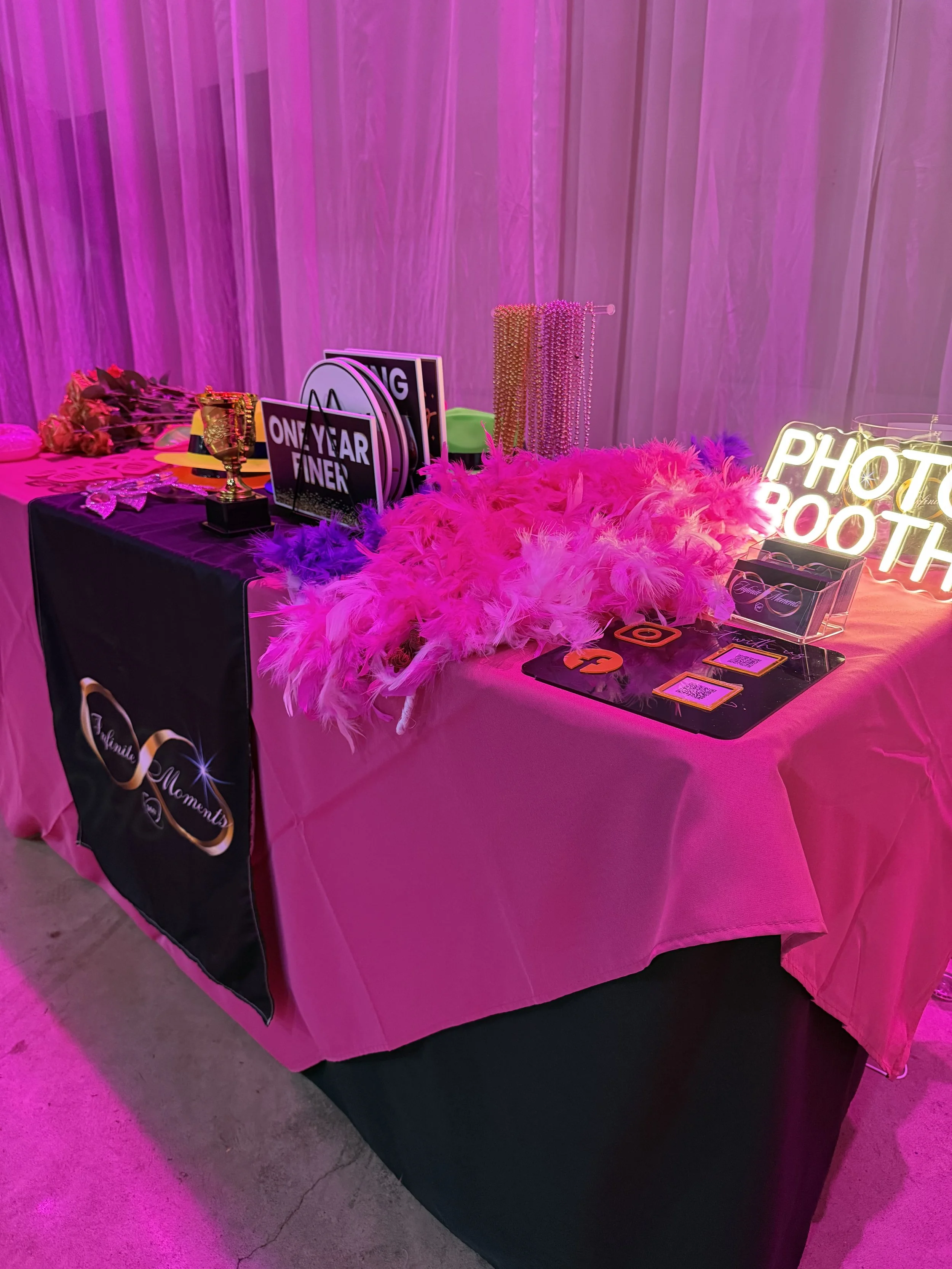 Photo booth table decorated with pink feathers, photo props, and neon sign with the text 'Photo Booth' at an event.