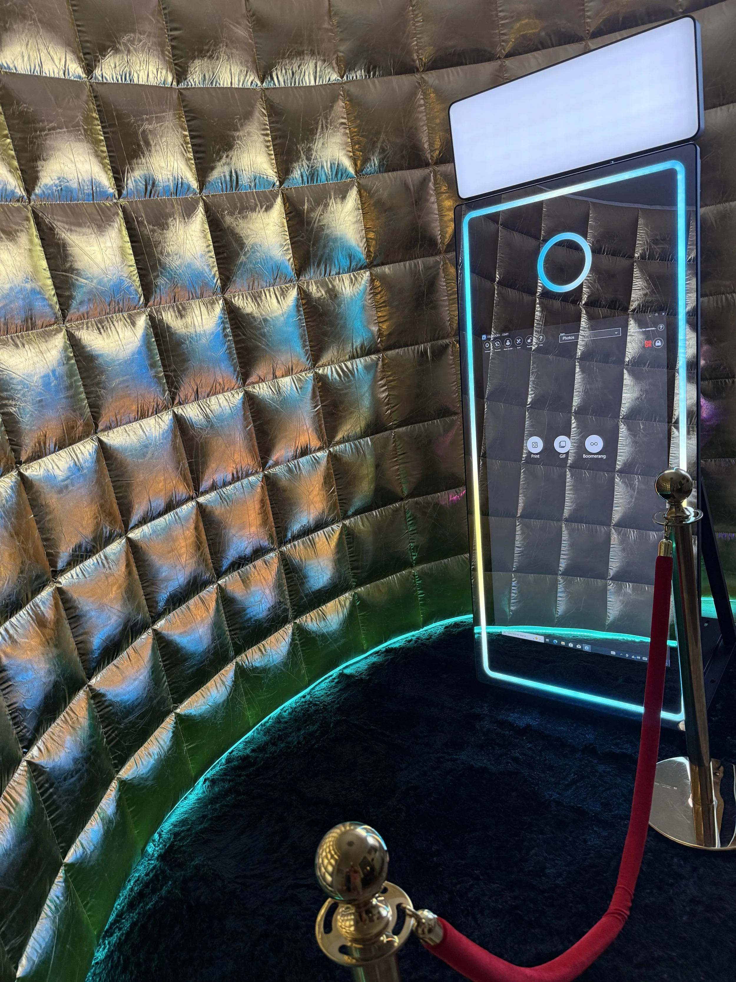 A photo booth with a reflective, padded gold wall background, a large mirror with neon lighting, and a red velvet rope barrier, used for photos or selfies.