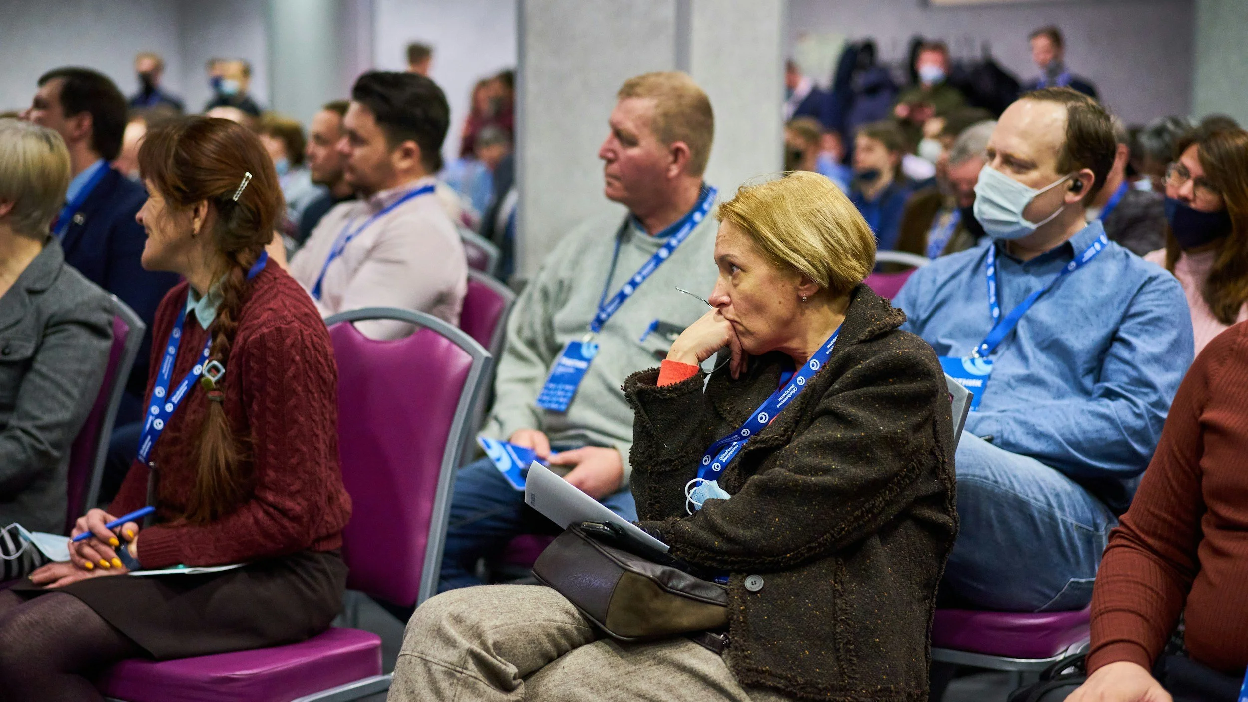 Audience seated at conference, wearing masks, listening attentively.
