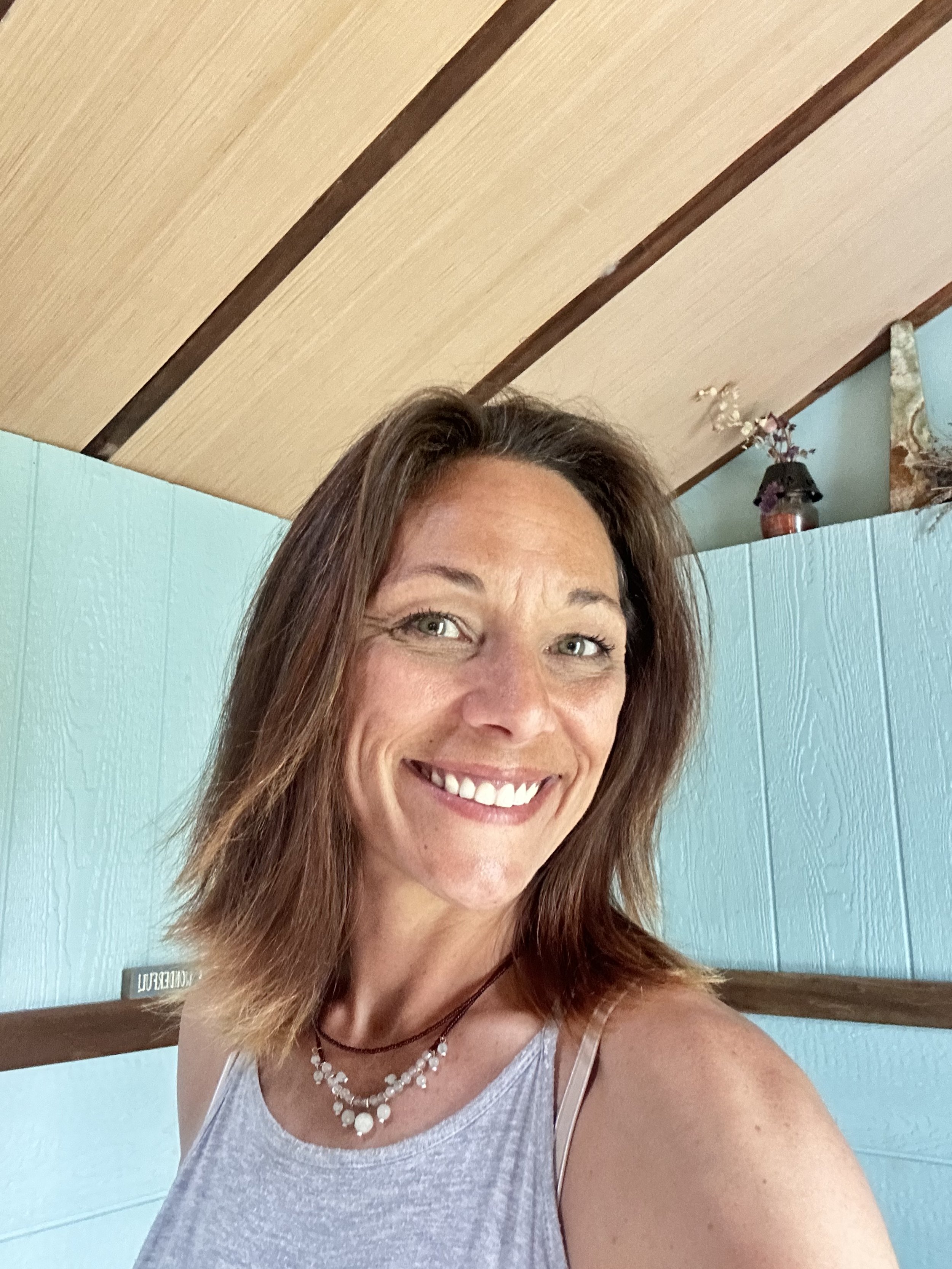 A woman smiling, wearing a gray tank top and a necklace, in a room with a light blue wall and a wooden ceiling.