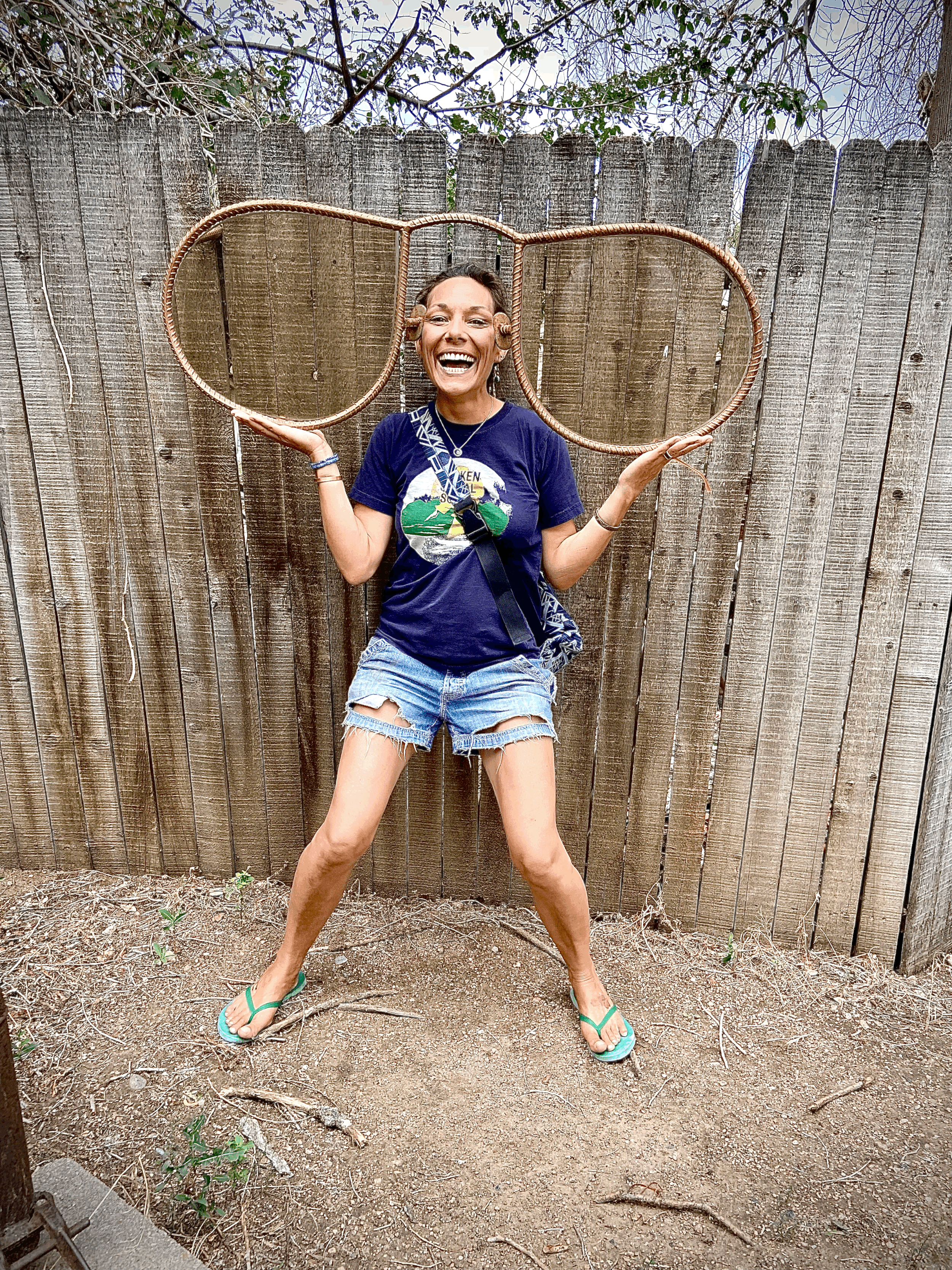 Woman smiling and holding oversized sunglasses frame in front of a wooden fence.