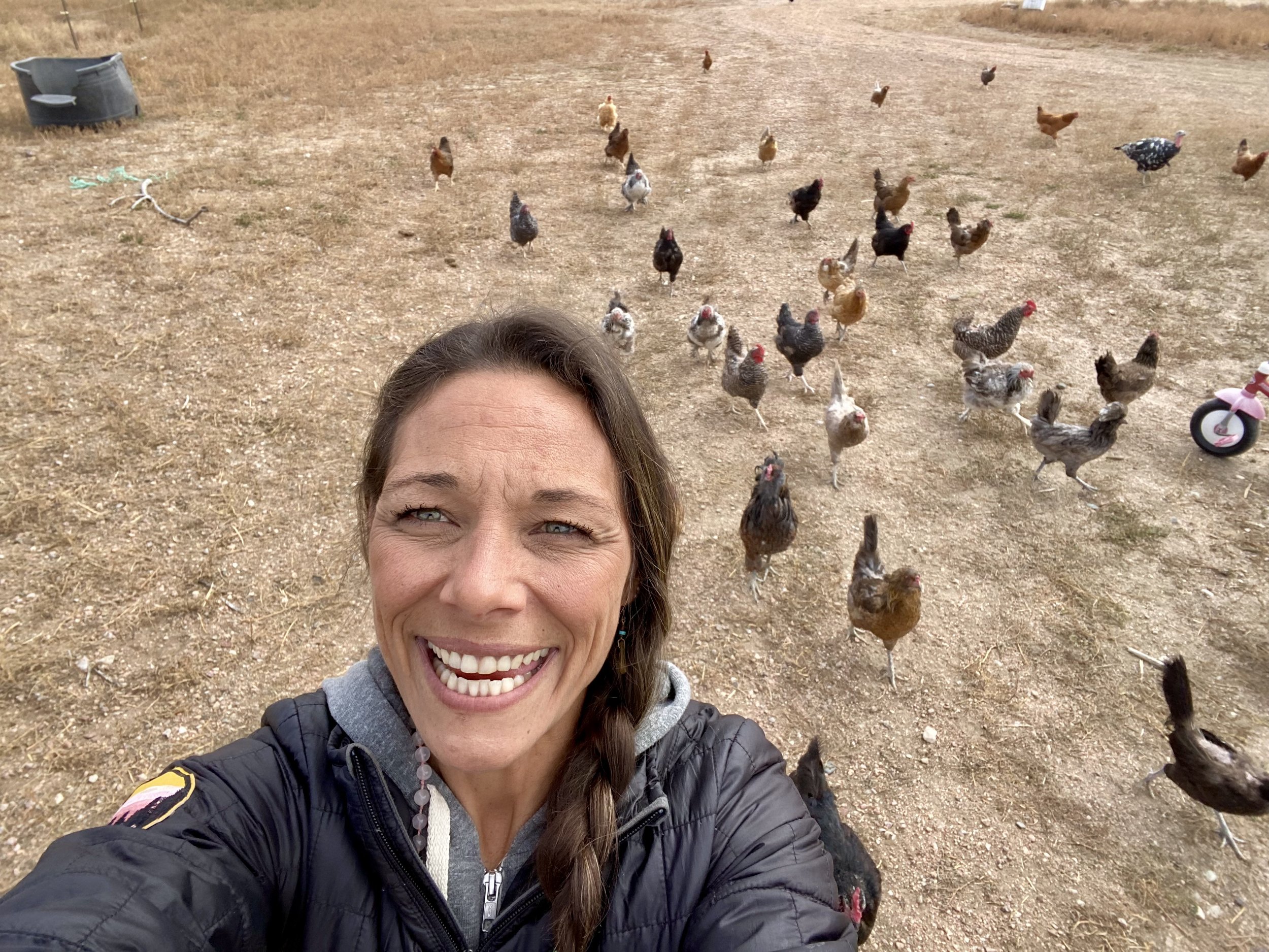 A woman taking a selfie outdoors on a dirt field with several chickens in the background.