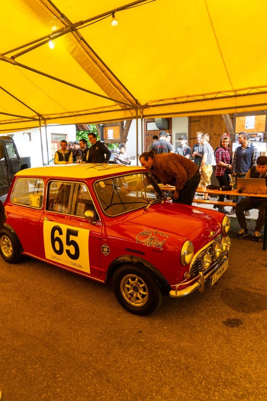 Auto da corsa rossa con il numero 65 all'interno di un tendone, con persone intorno che osservano e lavorano sulla macchina, ambientazione di un evento automobilistico.