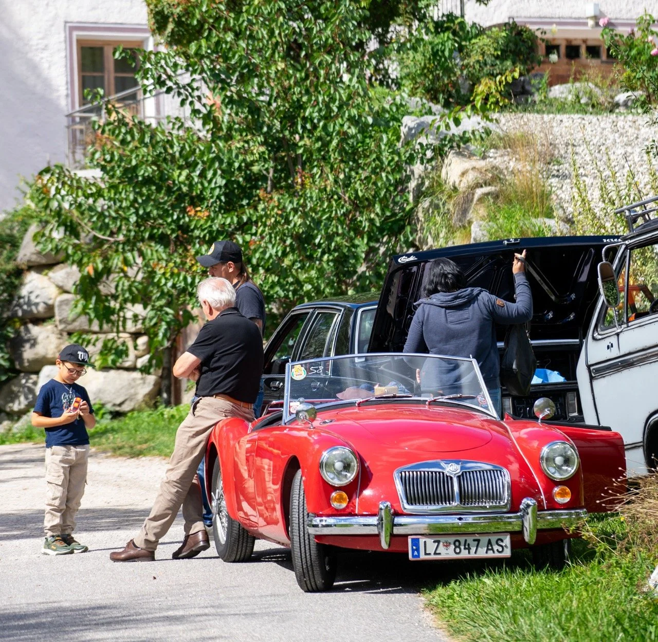 Cinque persone intorno a una vettura sportiva rossa con targa italiana e un veicolo bianco che mostra il bagagliaio aperto, in un'area verde con alberi e piante.