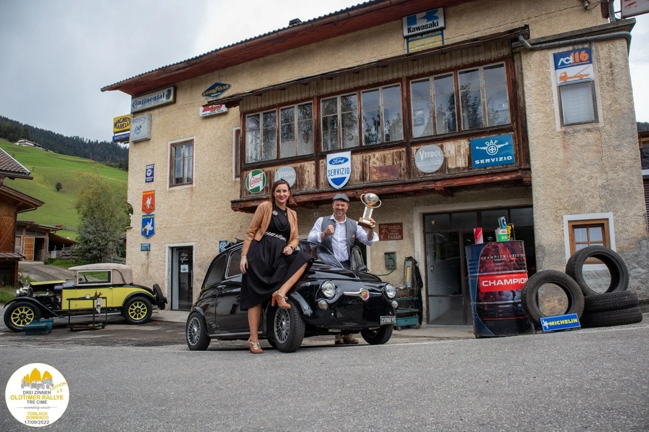 Una coppia festeggia davanti a un edificio con segnali di officina e un'auto vintage, un uomo tiene un trofeo e una donna si siede su un'auto, con uno sfondo di colline verdi.