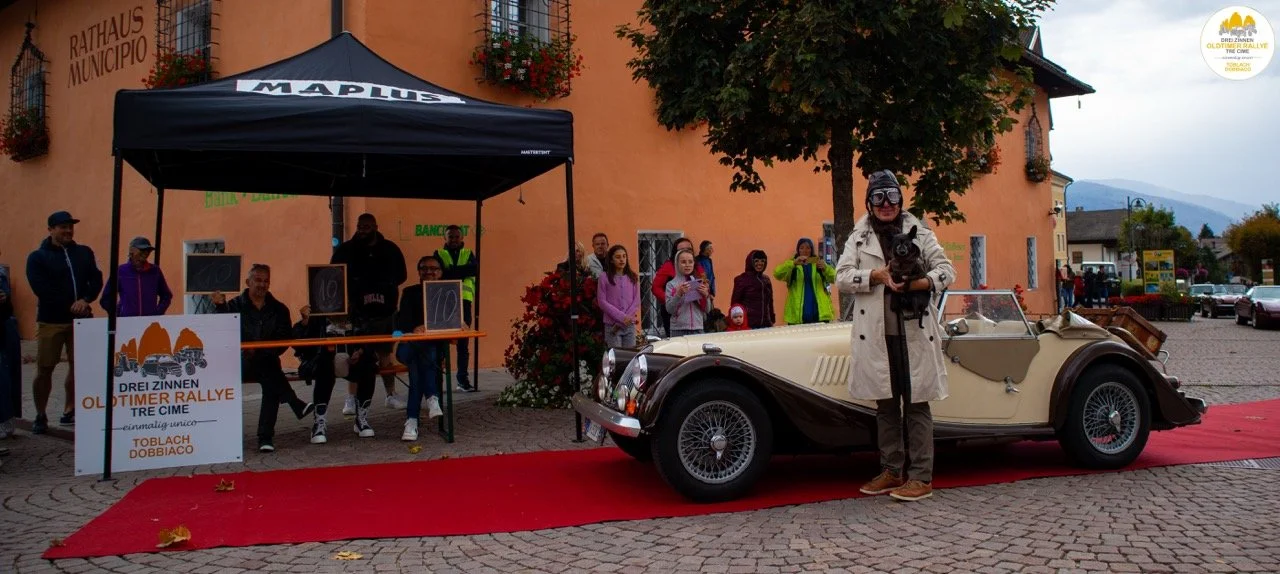 Una donna con occhiali e cappello posiziona un cane in un'auto d'epoca davanti a un municipio con una folla di persone che osservano, in una piazza con tappeto rosso, durante un evento ufficiale.
