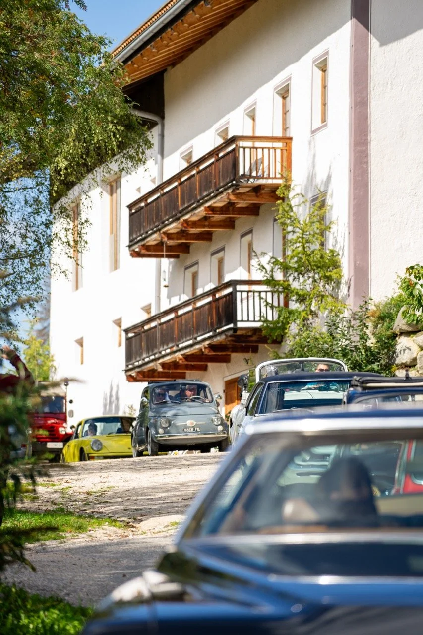 Auto d'epoca parcheggiate davanti a un edificio bianco con balconi di legno, circondate da vegetazione, in una giornata soleggiata.