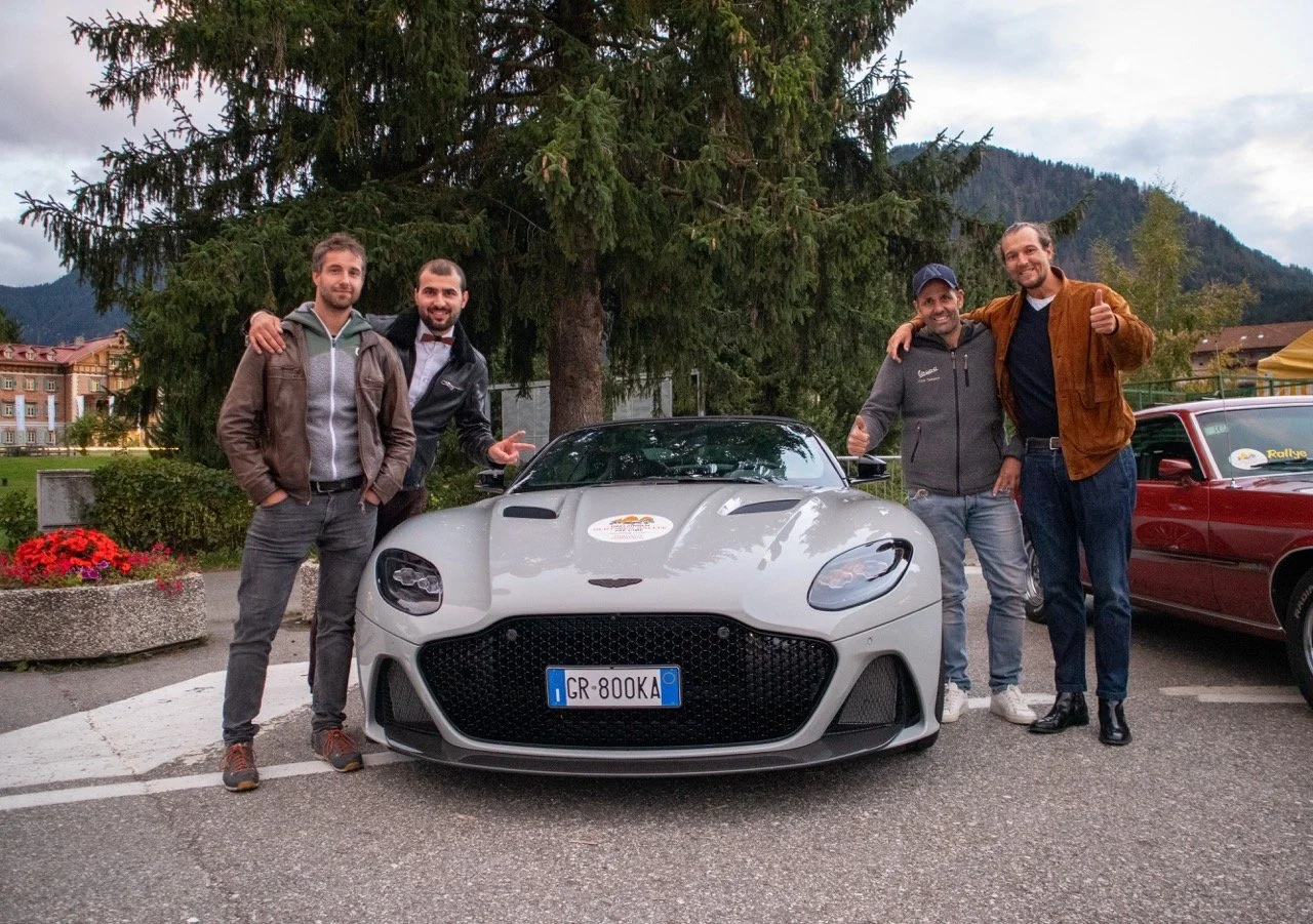 Cinque uomini sorridenti con abbigliamento casual posano insieme accanto a una moderna auto sportiva grigia con targa italiana, davanti a un albero e a un paesaggio montano.