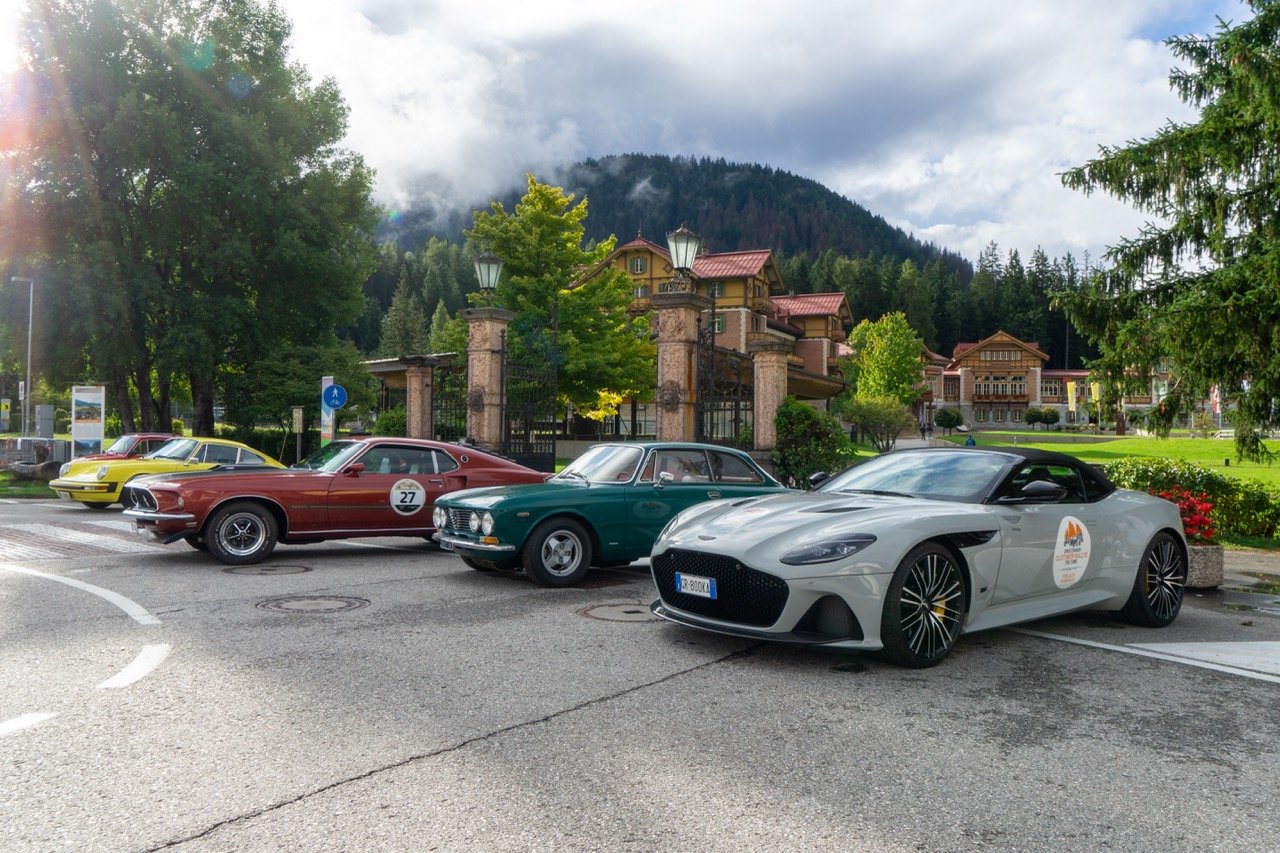 Auto sportiva moderna, auto classica, auto d'epoca e auto sportiva in un parcheggio con elementi naturali e case di montagna sullo sfondo.