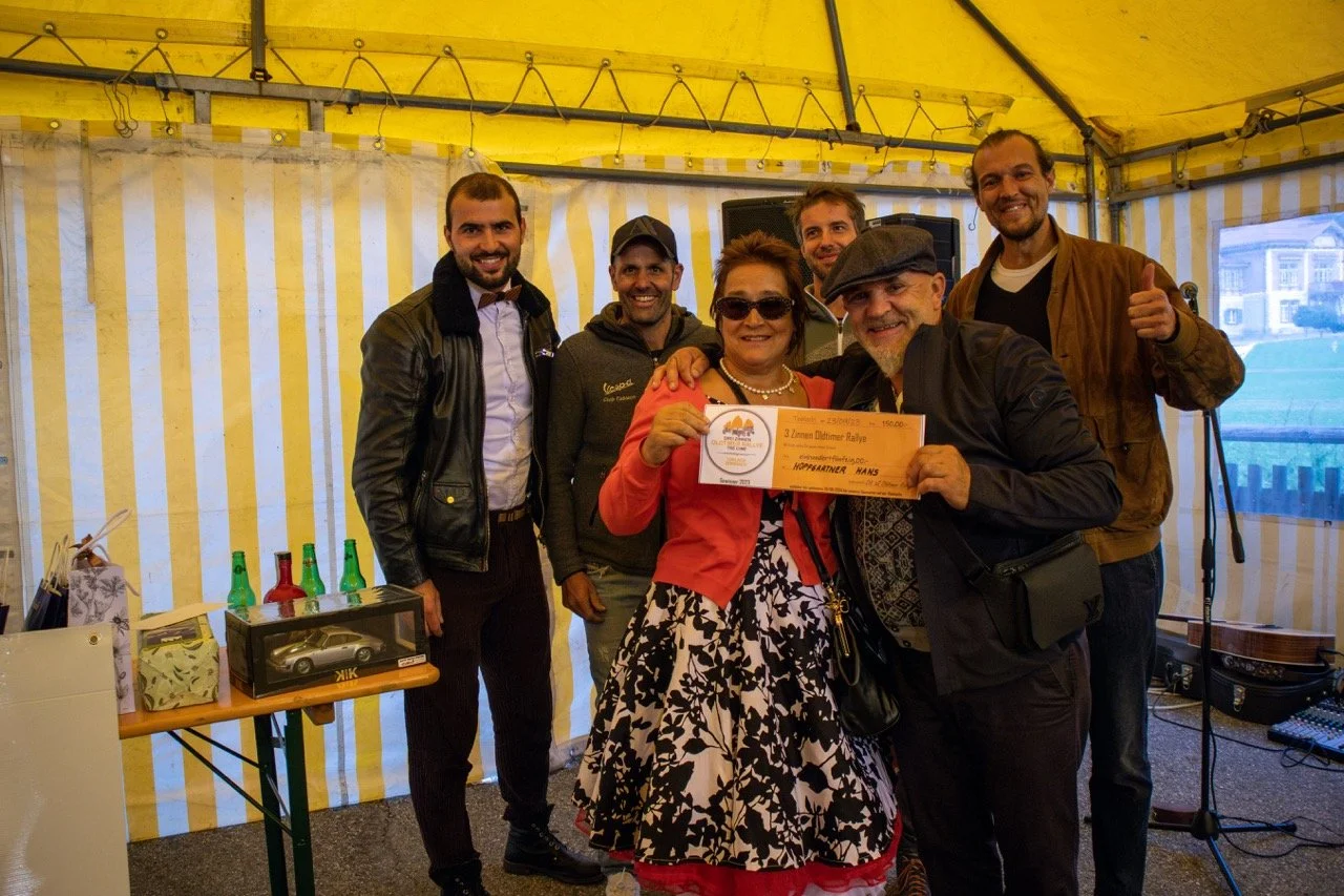 Gruppo di sette persone sorridenti sotto una tenda gialla, alcune che tengono un certificato o premio, con bottiglie colorate sul tavolo accanto.