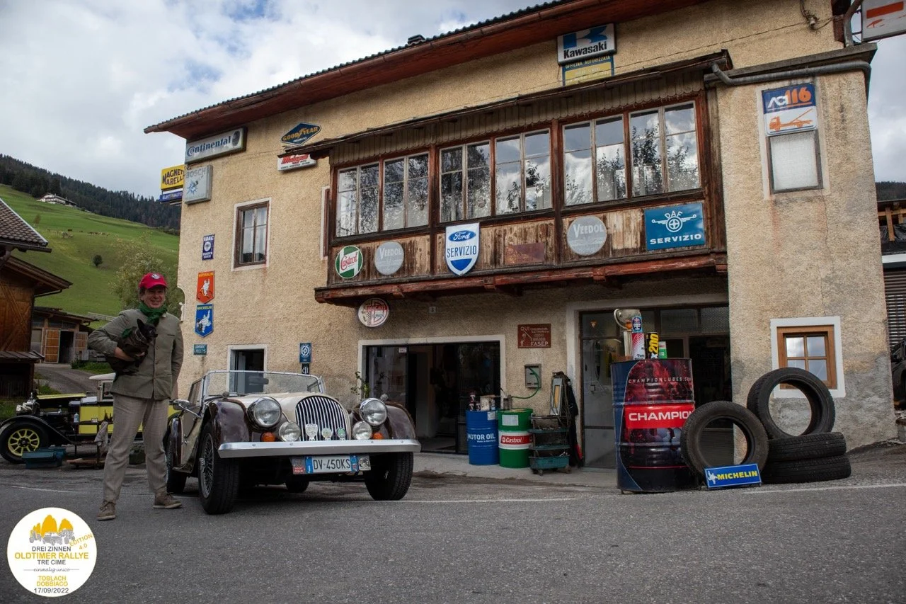 Una persona sorridente posa accanto a una macchina d'epoca davanti a un edificio in stile montano, con vari cartelli pubblicitari di marchi automobilistici e pneumatici, e alcuni pneumatici impilati. Sulla sinistra si vedono altri veicoli d'epoca.