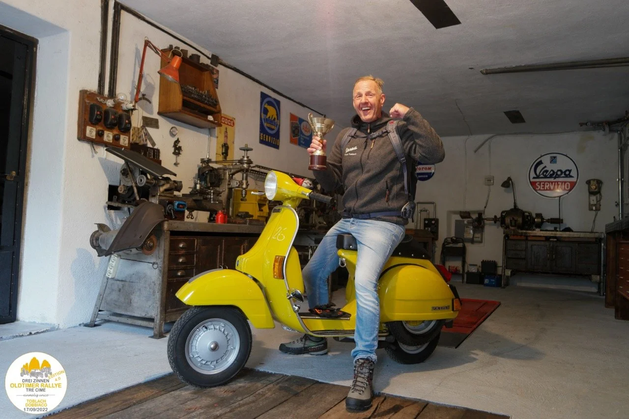 Uomo felice seduto su una Vespa gialla in un garage, che tiene in mano un trofeo e fa un pugno di gioia.