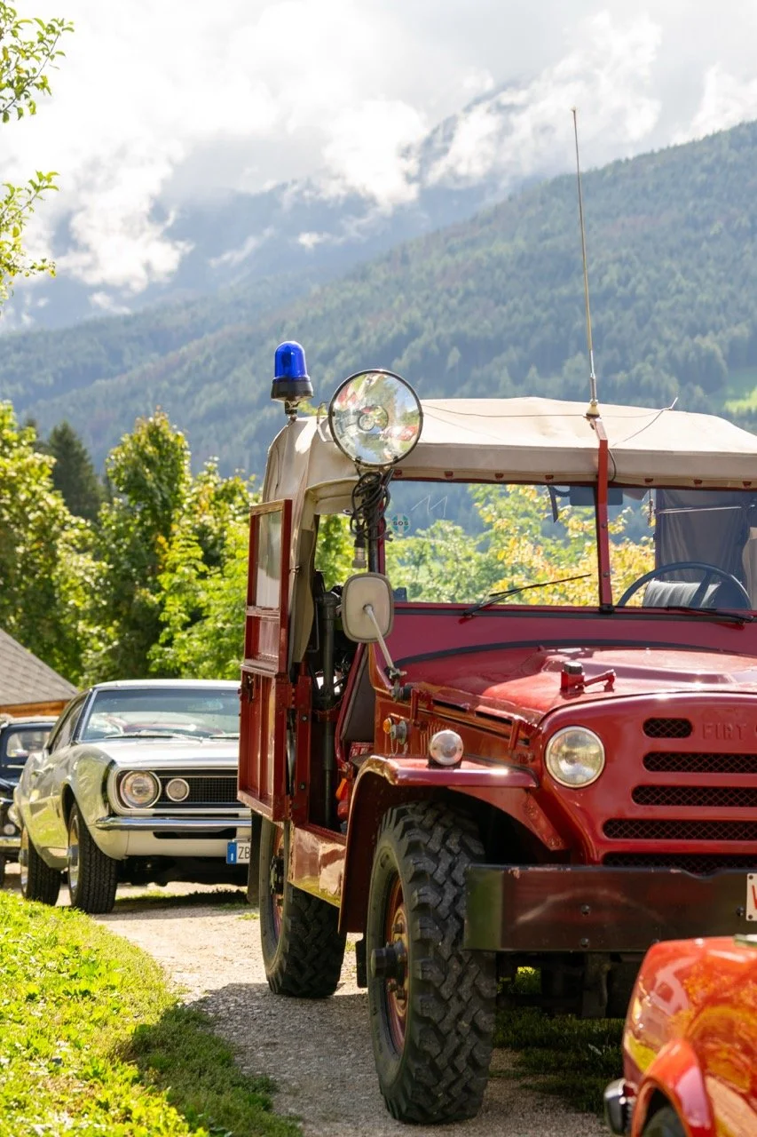 Veicolo da trasporto rosso in stile vintage con cappotta e luci lampeggianti blu e di riserva, parcheggiato tra altre auto nel contesto di un'area verde con montagne sullo sfondo.