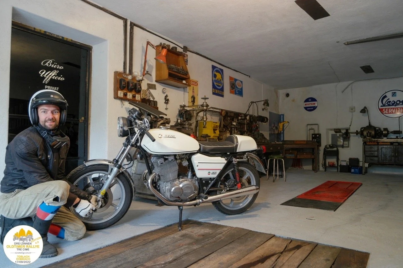 Uomo in tuta da motociclista e casco nero, che si trova accanto a una motocicletta bianca in un garage o officina.