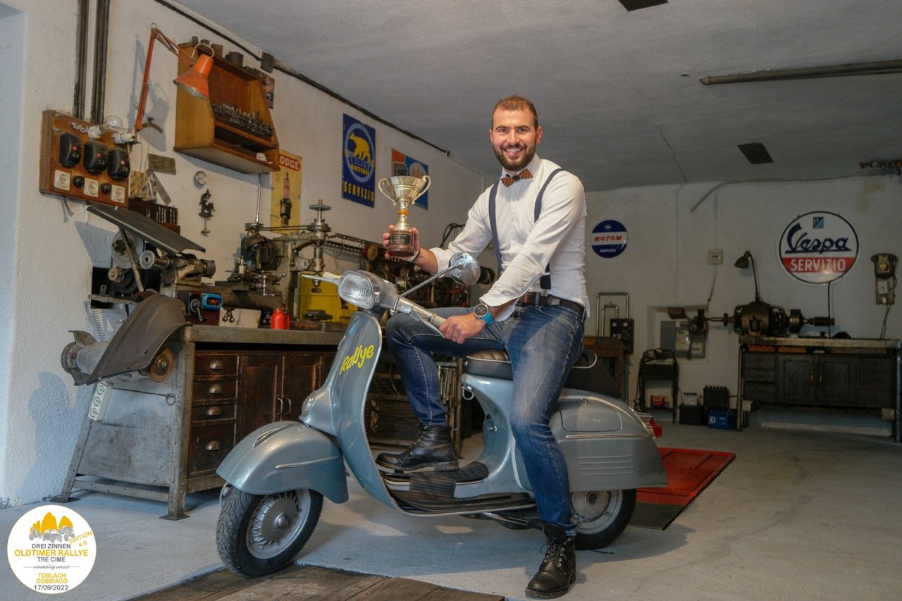 Uomo seduto su uno scooter vintage in un garage, sorride e tiene in mano un trofeo.