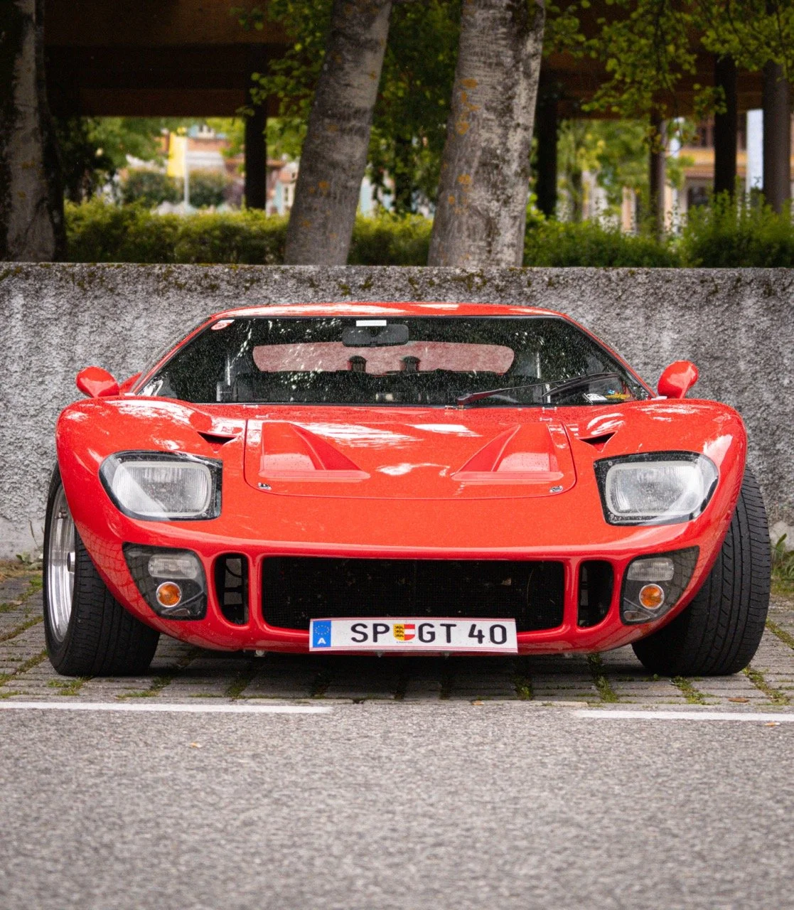 Auto sportiva di colore rosso parcheggiata su una strada di fronte a un muro di pietra, con alberi sullo sfondo.