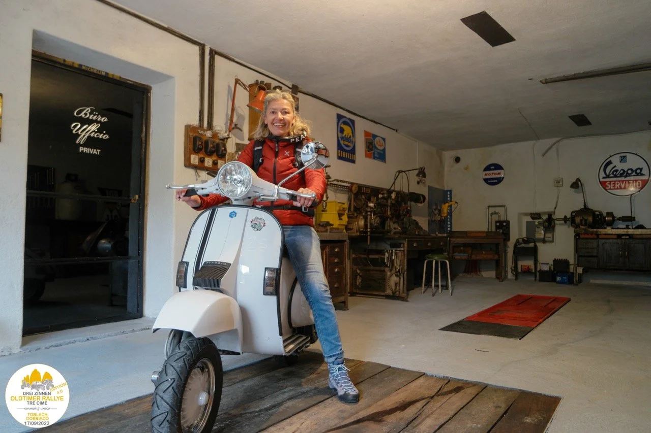Una donna sorride seduta su una vespa bianca dentro un garage o officina.