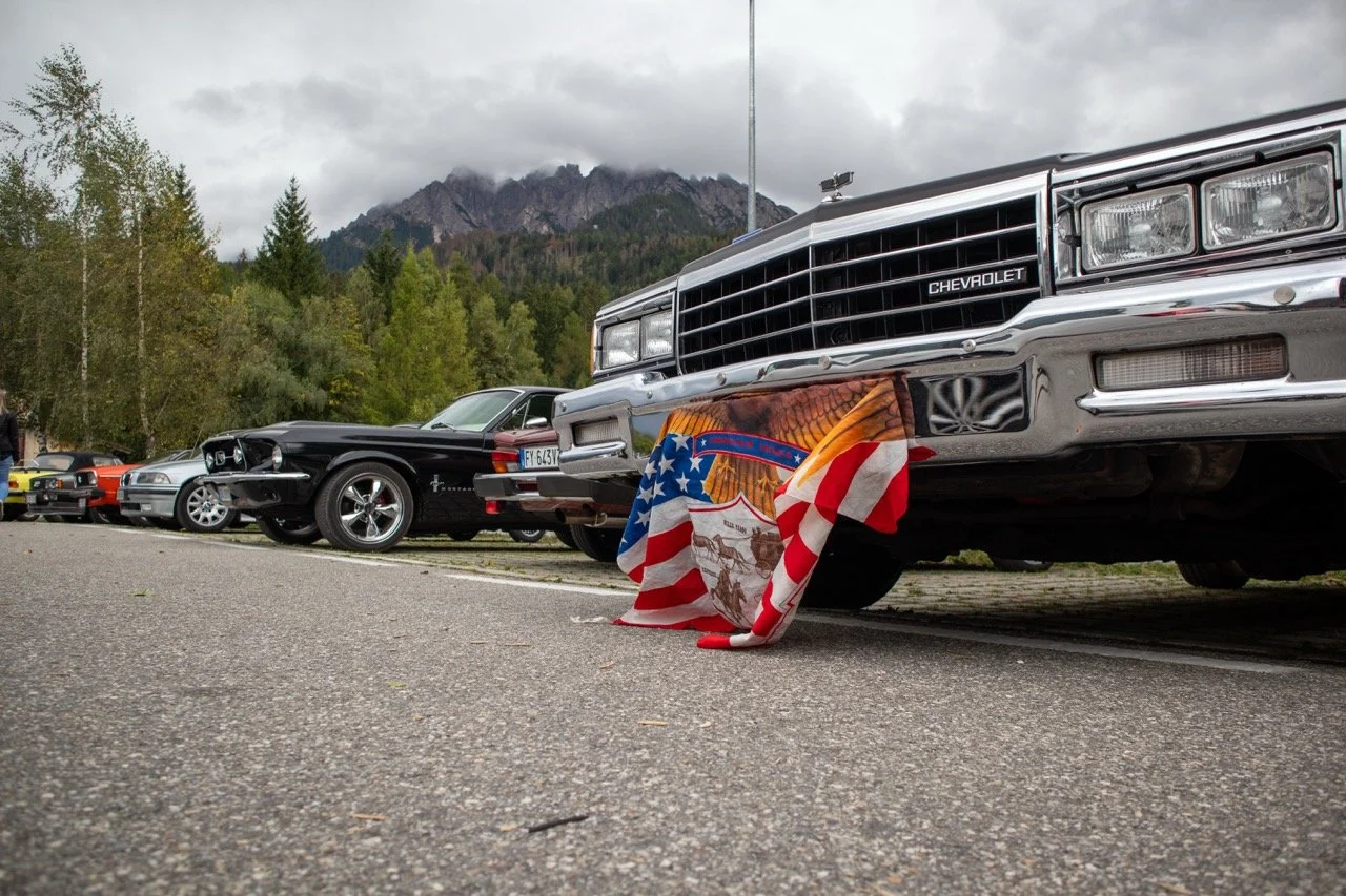 Auto d'epoca Chevrolet con bandiera americana come decorazione, circondato da altre automobili in un parcheggio con montagne sullo sfondo.