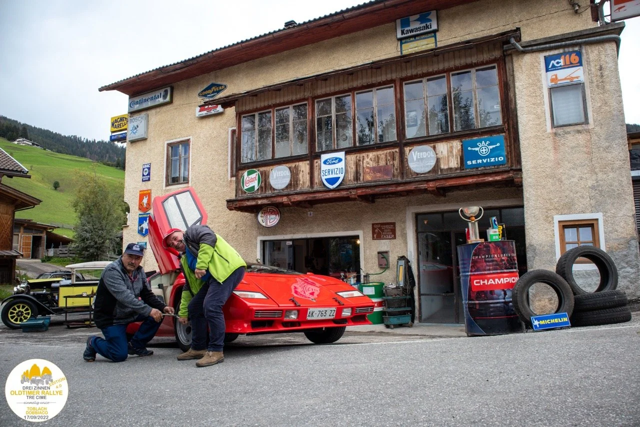Due uomini si occupano di un'auto sportiva rossa con motore sollevato, davanti a un edificio con vari cartelli pubblicitari e gomme usate, in un contesto montano.