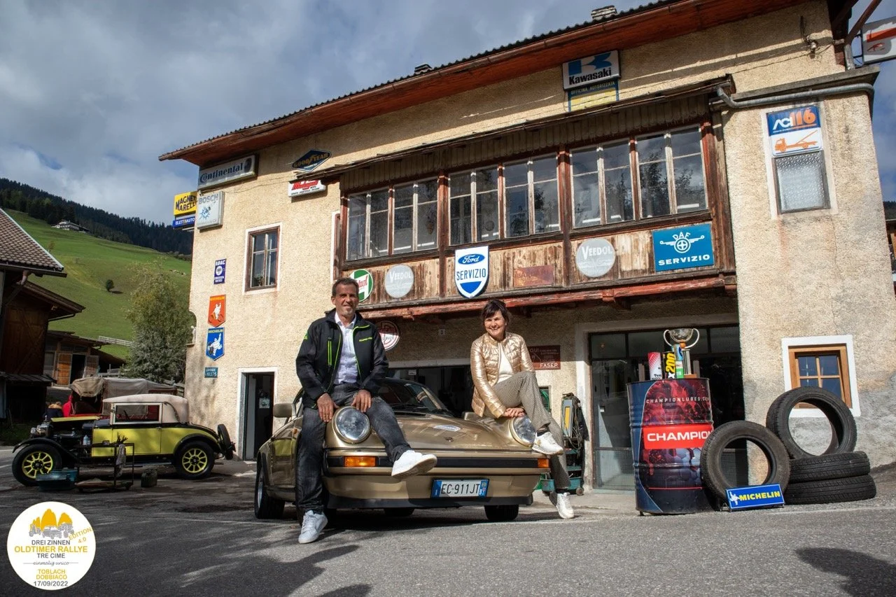 Due persone sorridenti si trovano davanti a un edificio con numerosi cartelli di officina e auto d'epoca, una delle quali è una Porsche 911, in un contesto di montagna con altri veicoli e attrezzature da officina