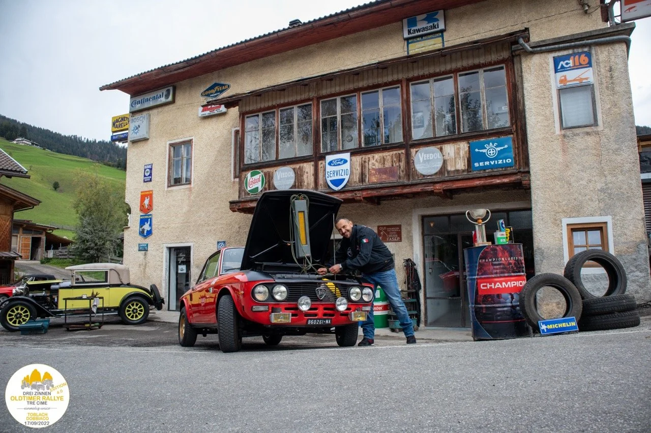 Un uomo apre il cofano di una vettura d'epoca rossa davanti a un edificio con vari segni e loghi di aziende di automobili e pneumatici. Sul lato destro, ci sono pneumatici e un caricatore di trofei.