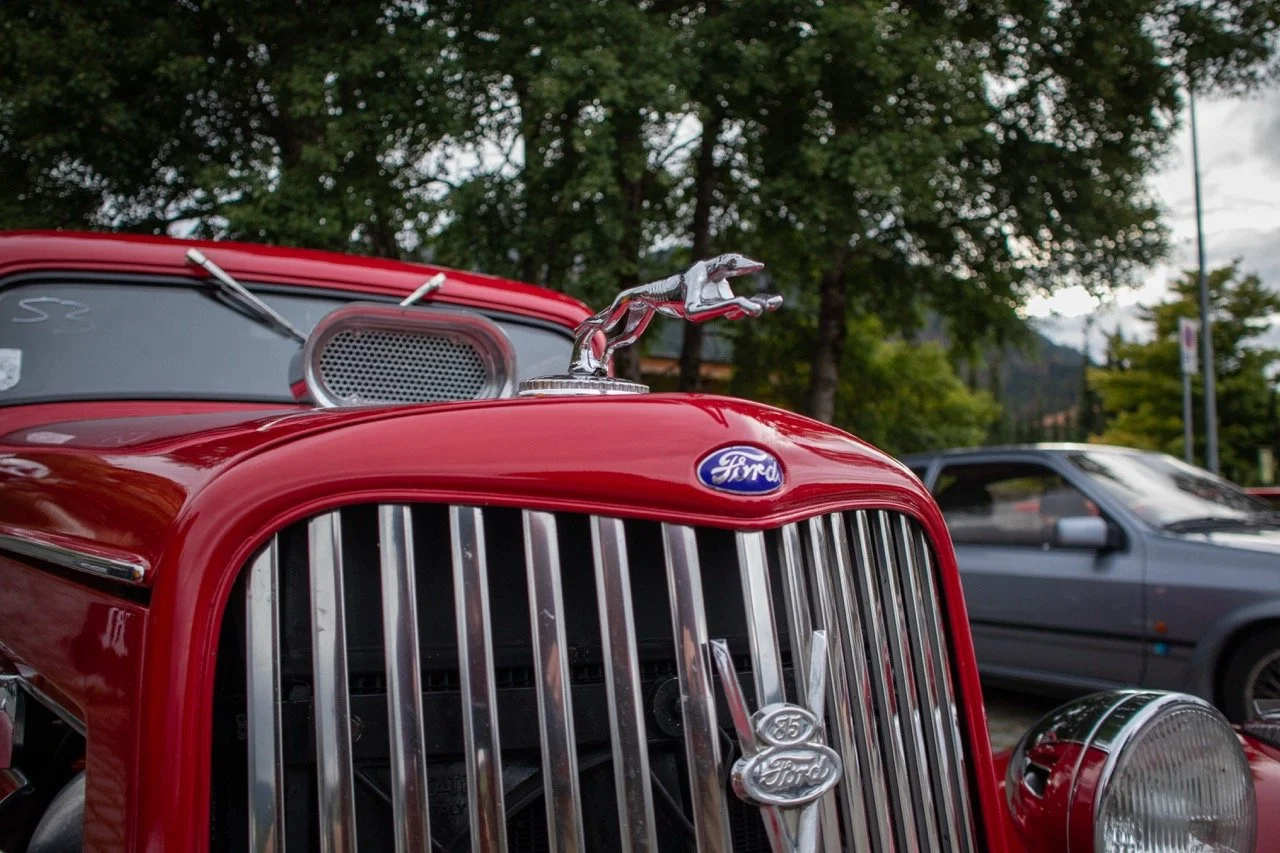 Dettaglio di un'auto d'epoca rossa, con griglia frontale cromata, logo Ford e ornamento a forma di cavallo al centro del cofano, parcheggiata all'aperto con altri veicoli sullo sfondo e alberi verdi.