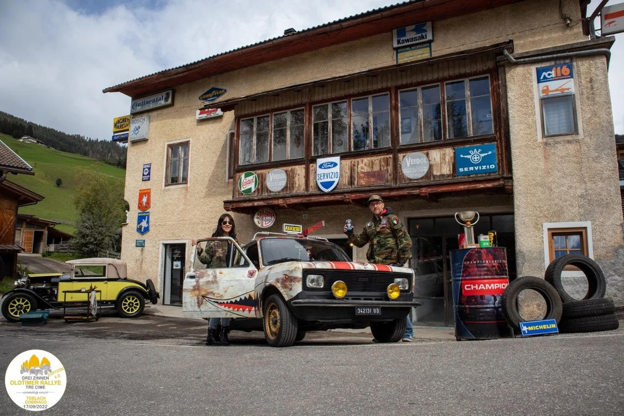 Due persone con abiti da rally posano con una macchina decorata a tema corsa in un garage di un'officina di auto da rally, con pneumatici usati e vari logo di marche di pneumatici e ricambi.