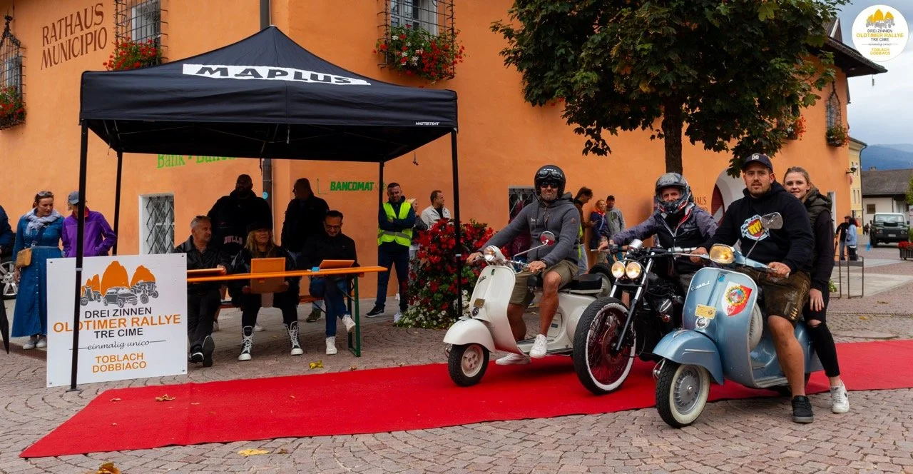 Gruppo di persone che partecipano a un rally old timer con moto e scooter d'epoca davanti al municipio, con un tappeto rosso e un'area di registrazione con persone sedute sotto un gazebo.