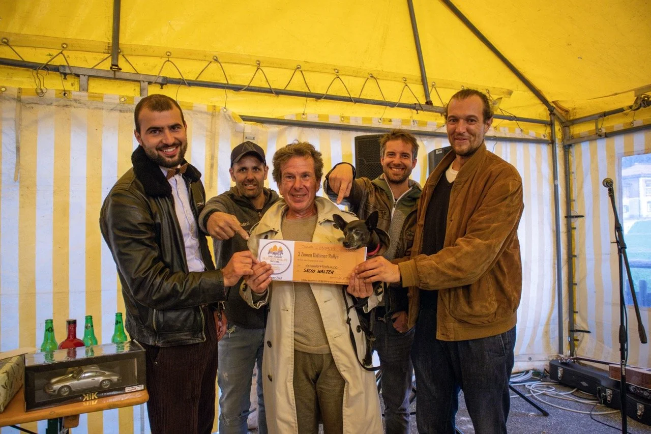 Gruppo di uomini sorridenti con un cane nero in un evento sotto una tenda gialla, uno di loro tiene un certificato di vincita.