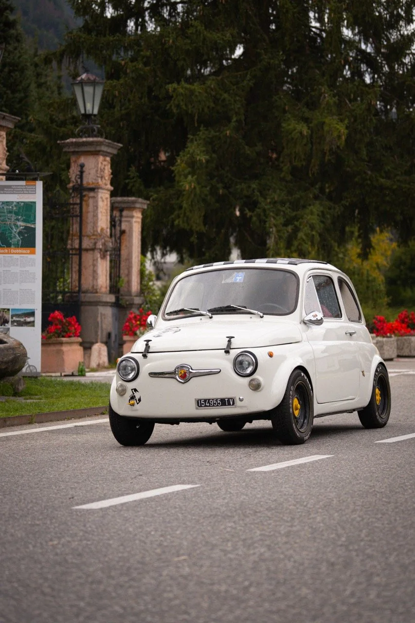 Una Fiat 500 d'epoca bianca parcheggiata lungo la strada, con sfondo di alberi e una cancellata in pietra.