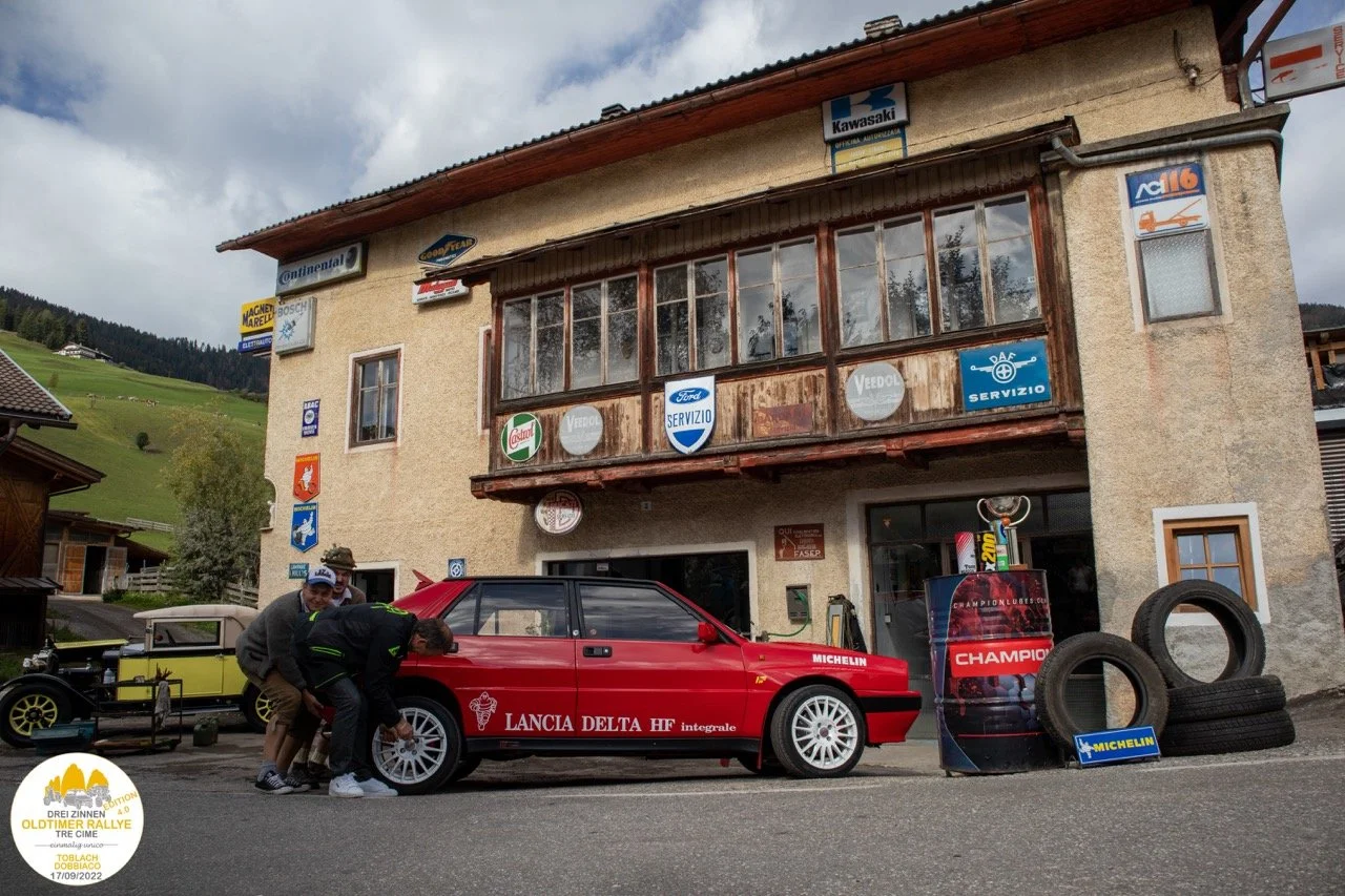 Auto da rally Lancia Delta HF integrale parcheggiata davanti a un edificio con molte pubblicità, due persone vicino all'auto, pneumatici e cartelloni pubblicitari in primo piano.